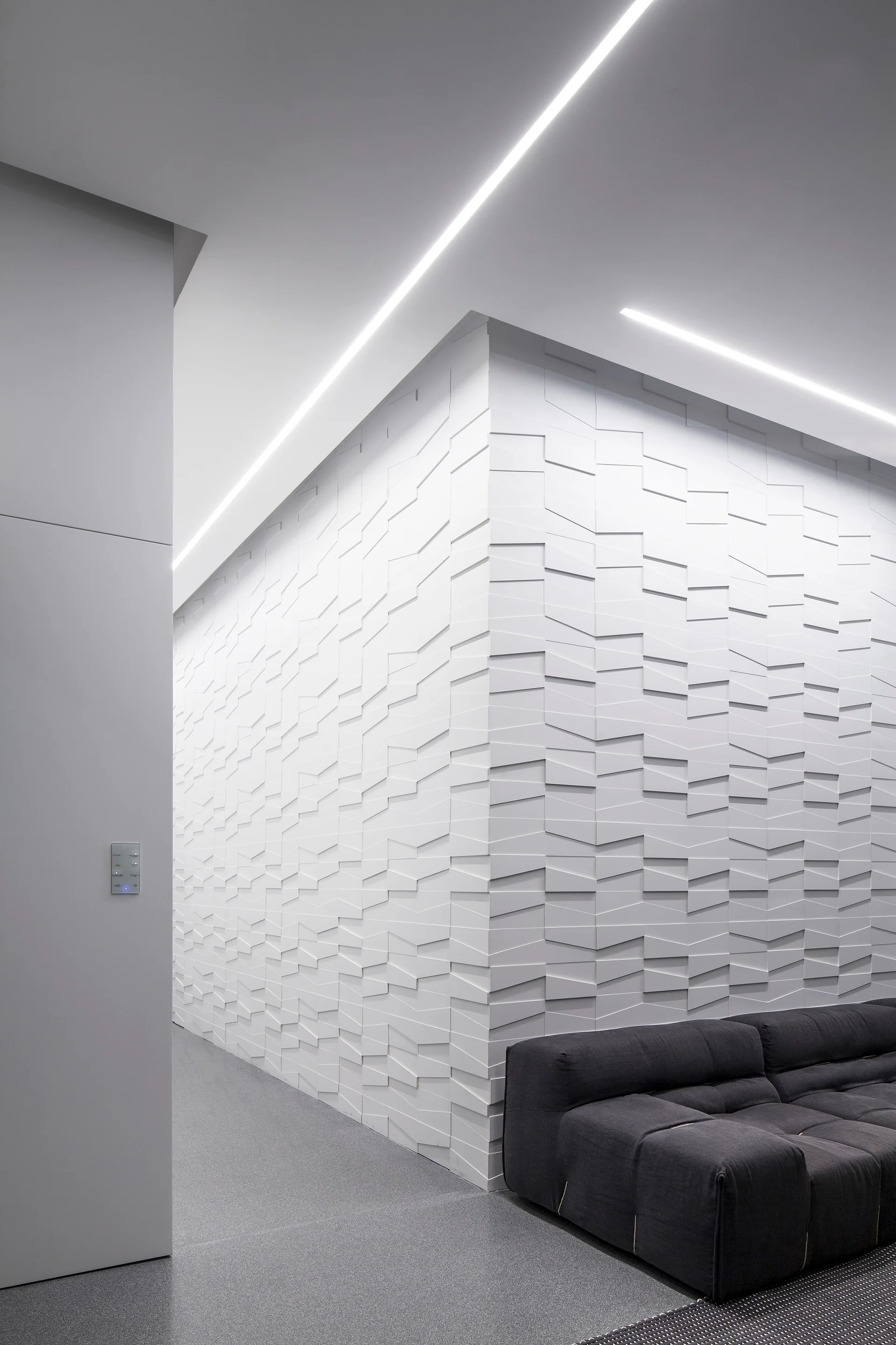A modern lounge features a dark grey low-profile sofa next to a white wall with geometric 3D paneling under linear ceiling lighting.