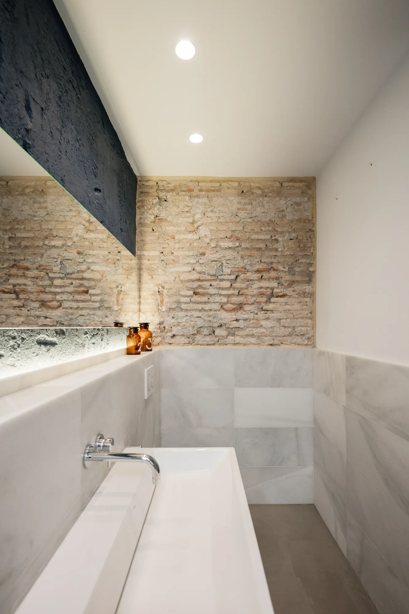 A modern bathroom features a white integrated sink with a wall-mounted chrome faucet beneath a large mirror and an exposed brick wall.