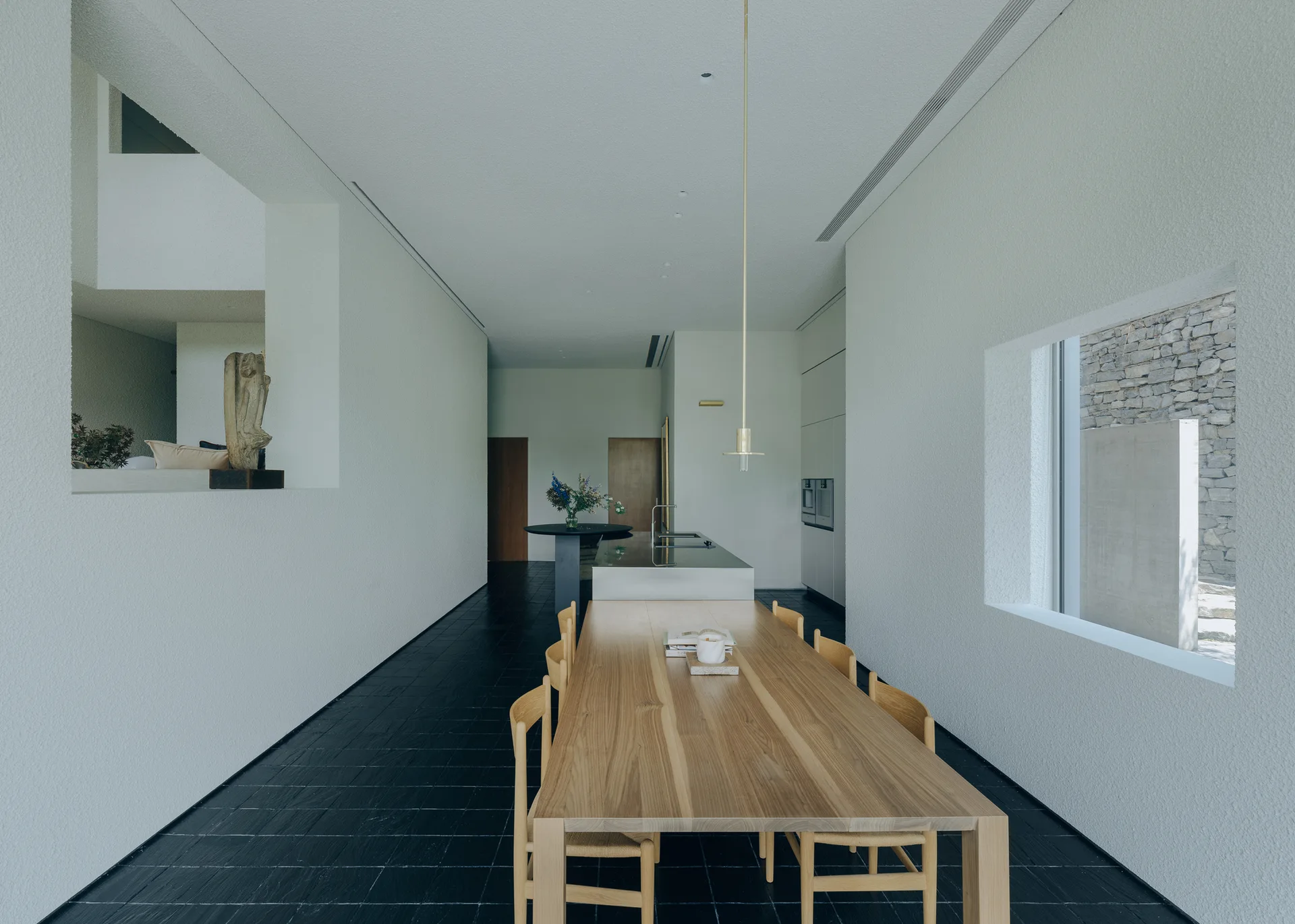 A long natural wood dining table and matching chairs sit on a dark stone tile floor in a minimalist white room with a kitchen island.