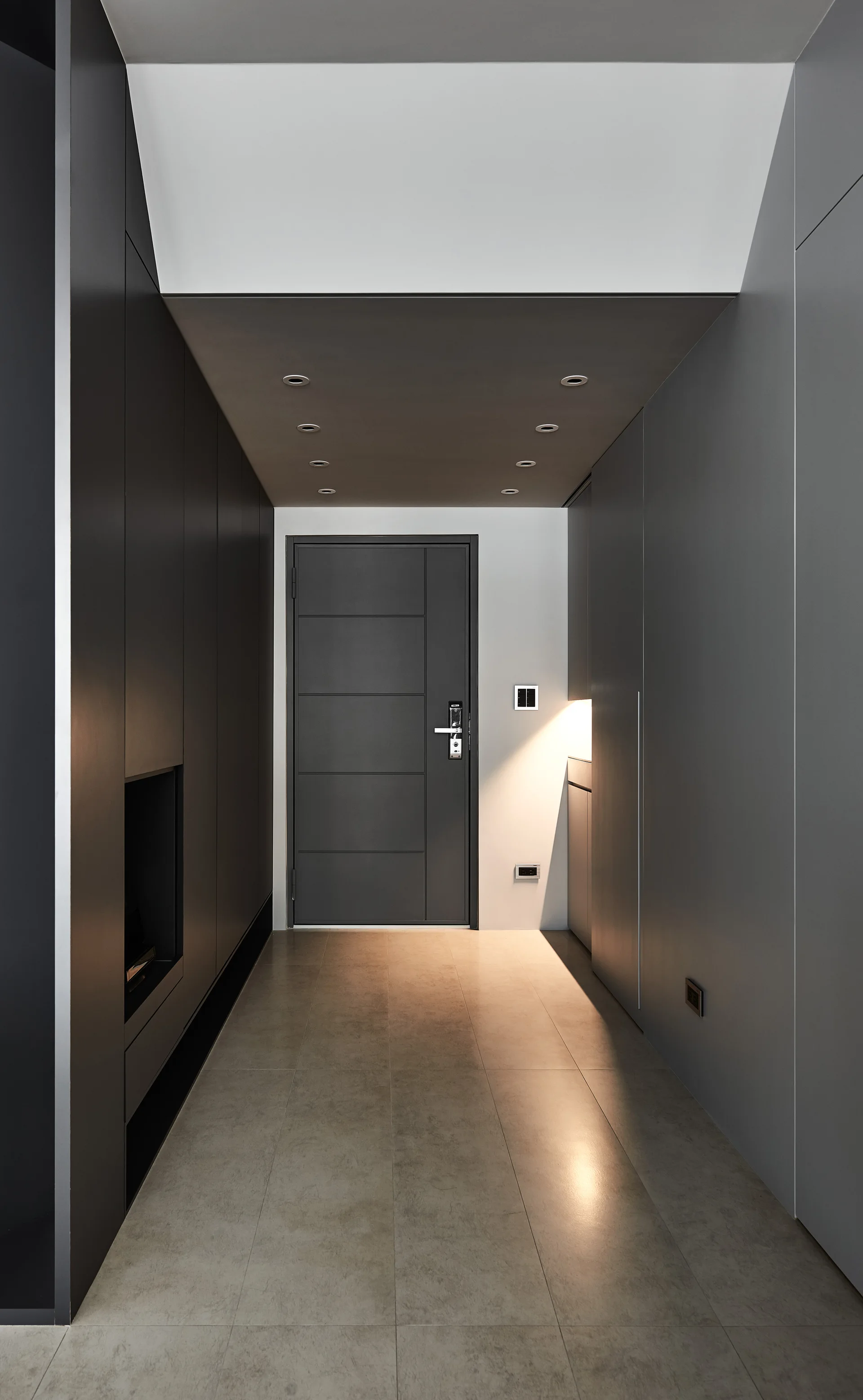 A modern entryway features charcoal grey built-in cabinets, a matching paneled door, large floor tiles, and recessed ceiling spotlights.