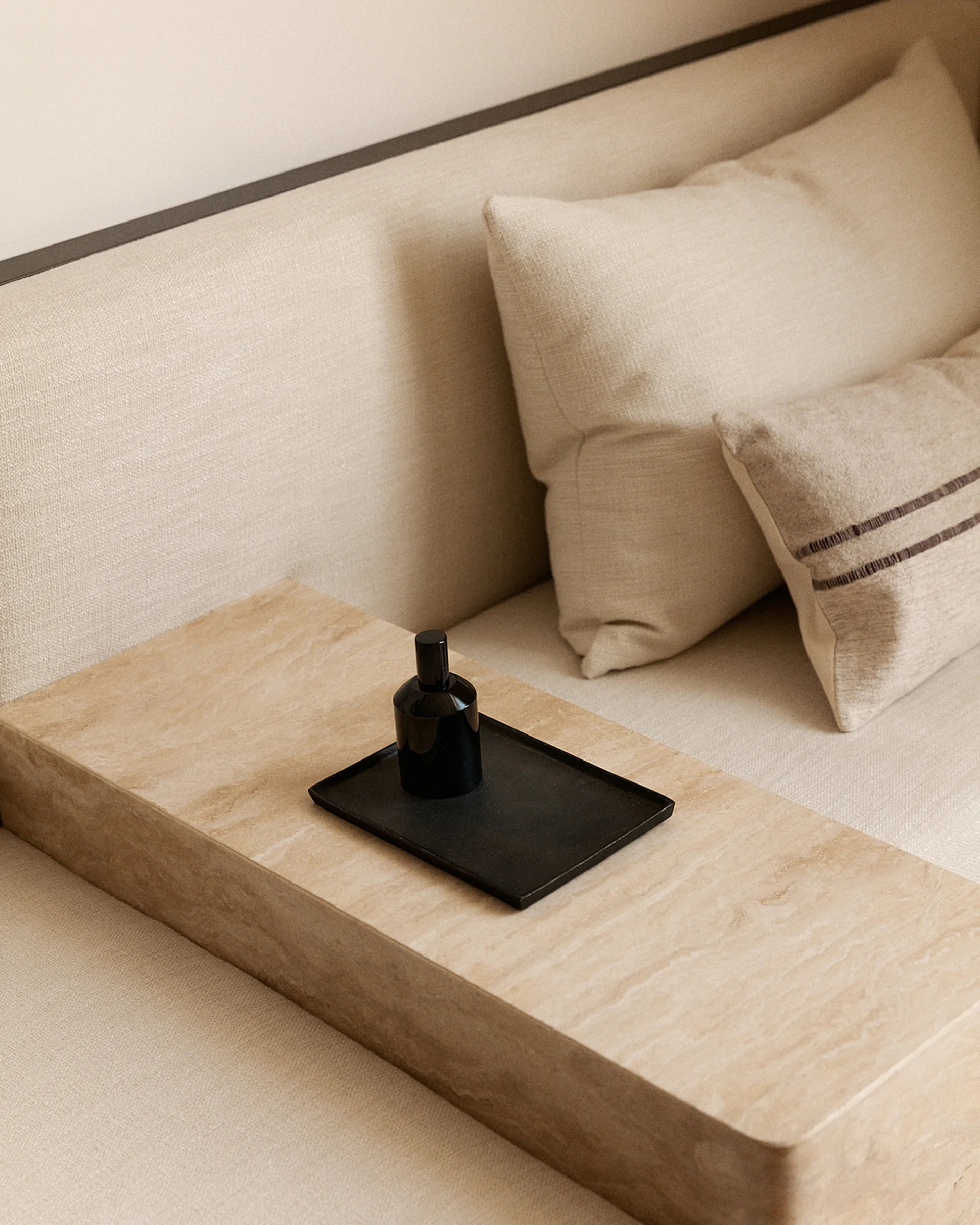 A close-up of a beige upholstered daybed featuring an integrated travertine stone ledge holding a small black tray and decorative bottle.