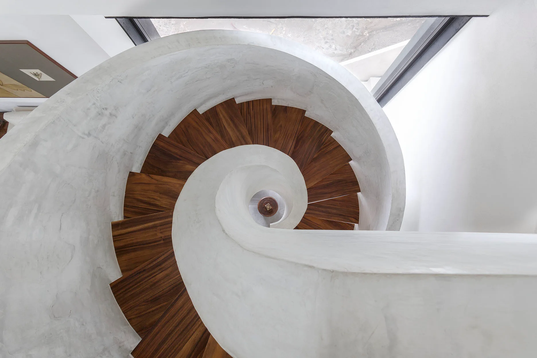 A top-down view of a helical spiral staircase featuring rich brown wooden treads and a smooth white plaster railing.