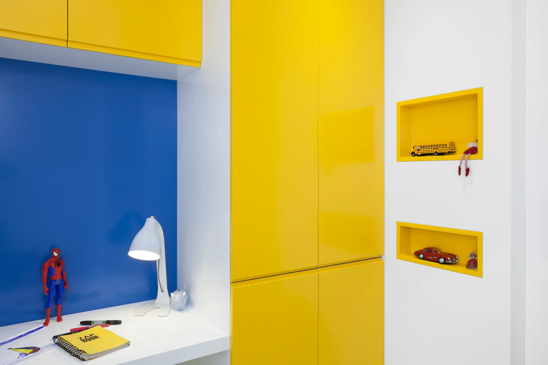 A modern kids' study area featuring bright yellow built-in cabinets, a white desk with a blue backdrop, and two yellow recessed wall niches.