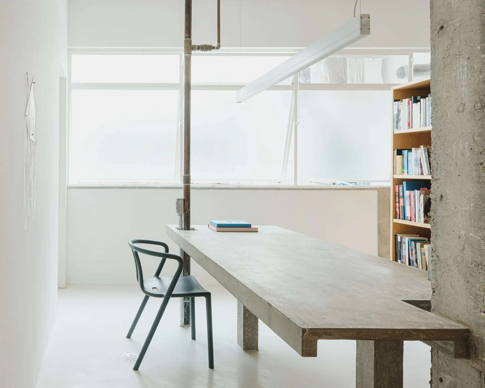 A minimalist home office featuring a long concrete slab desk, a black curved chair, an exposed metal pipe, and a tall wooden bookshelf.