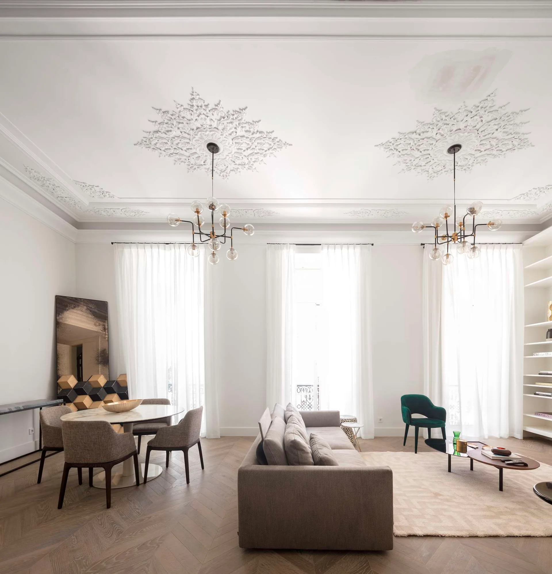 A bright living space features a gray sofa, marble dining table, and herringbone wood floors beneath detailed plaster ceiling medallions.