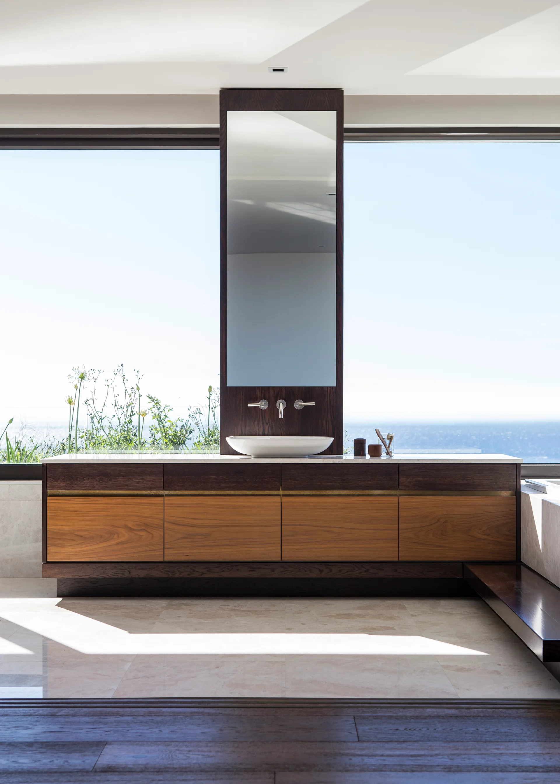 A minimalist bathroom features a floating wooden vanity with a vessel sink and a tall mirror column in front of a large window overlooking the ocean.