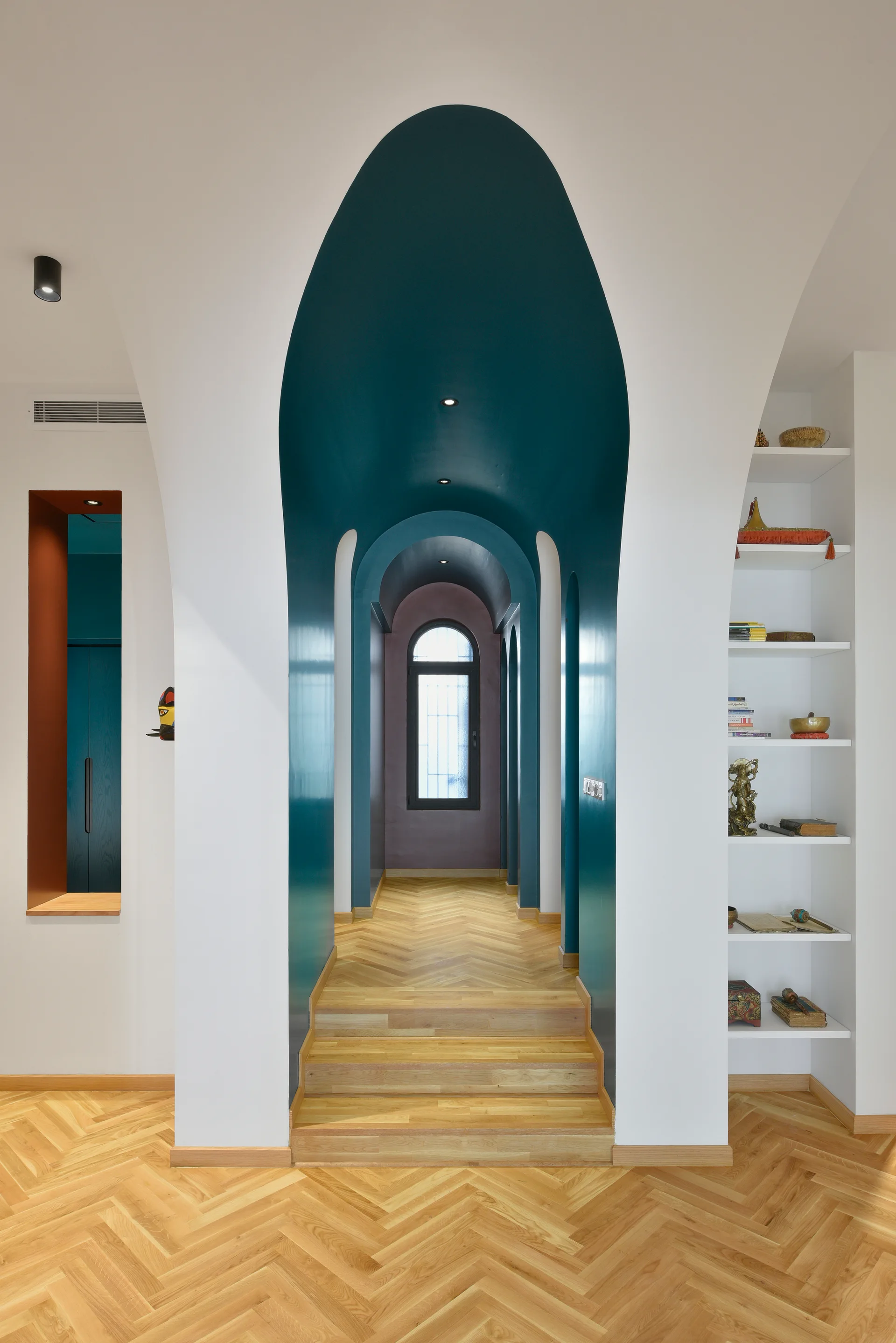 An interior hallway features nested teal-painted archways leading to an arched window, with white walls and herringbone wood flooring.