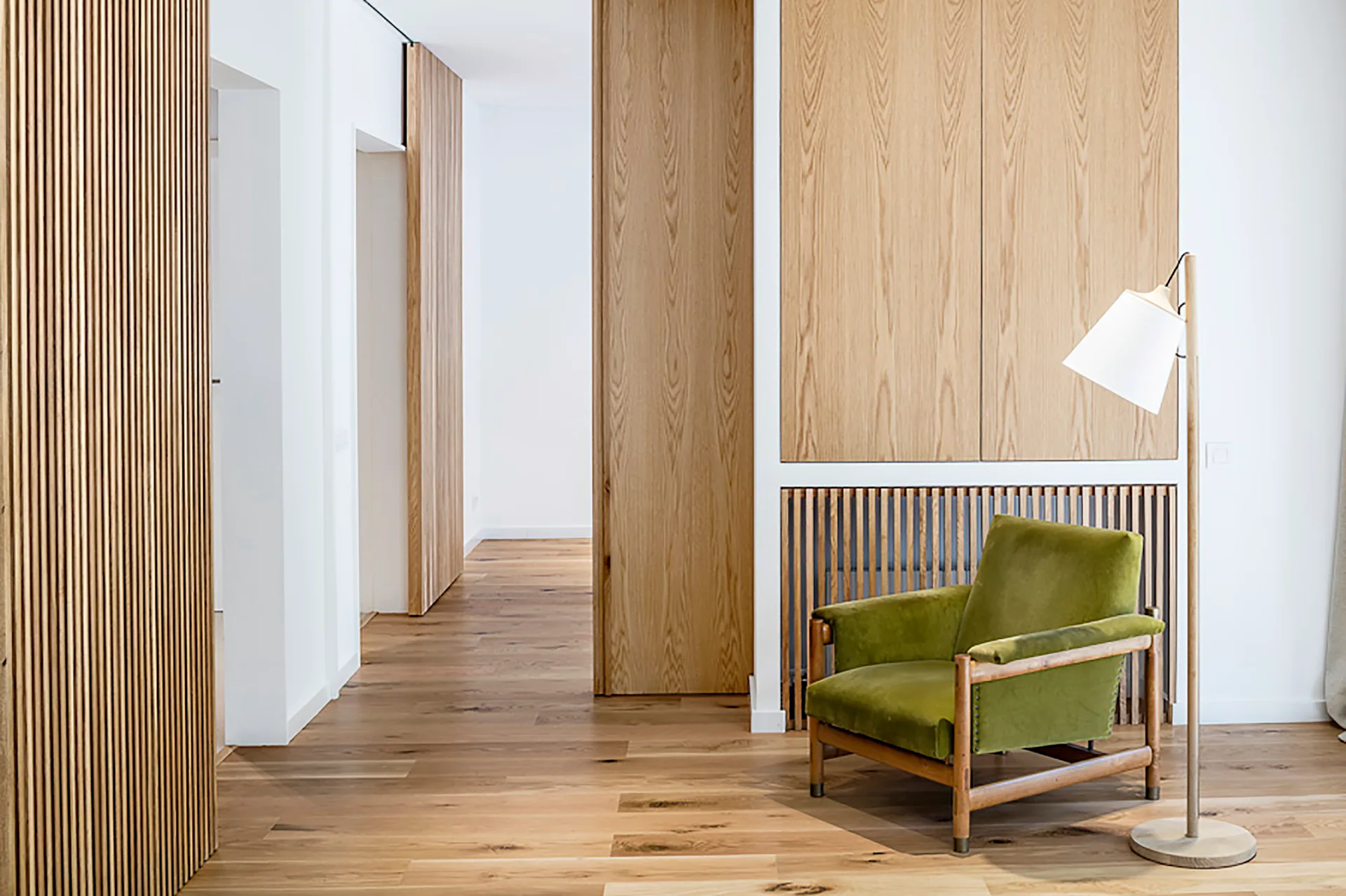 An olive green velvet armchair stands next to a wooden floor lamp in a hallway featuring light oak wood wall panels and vertical wood slats.