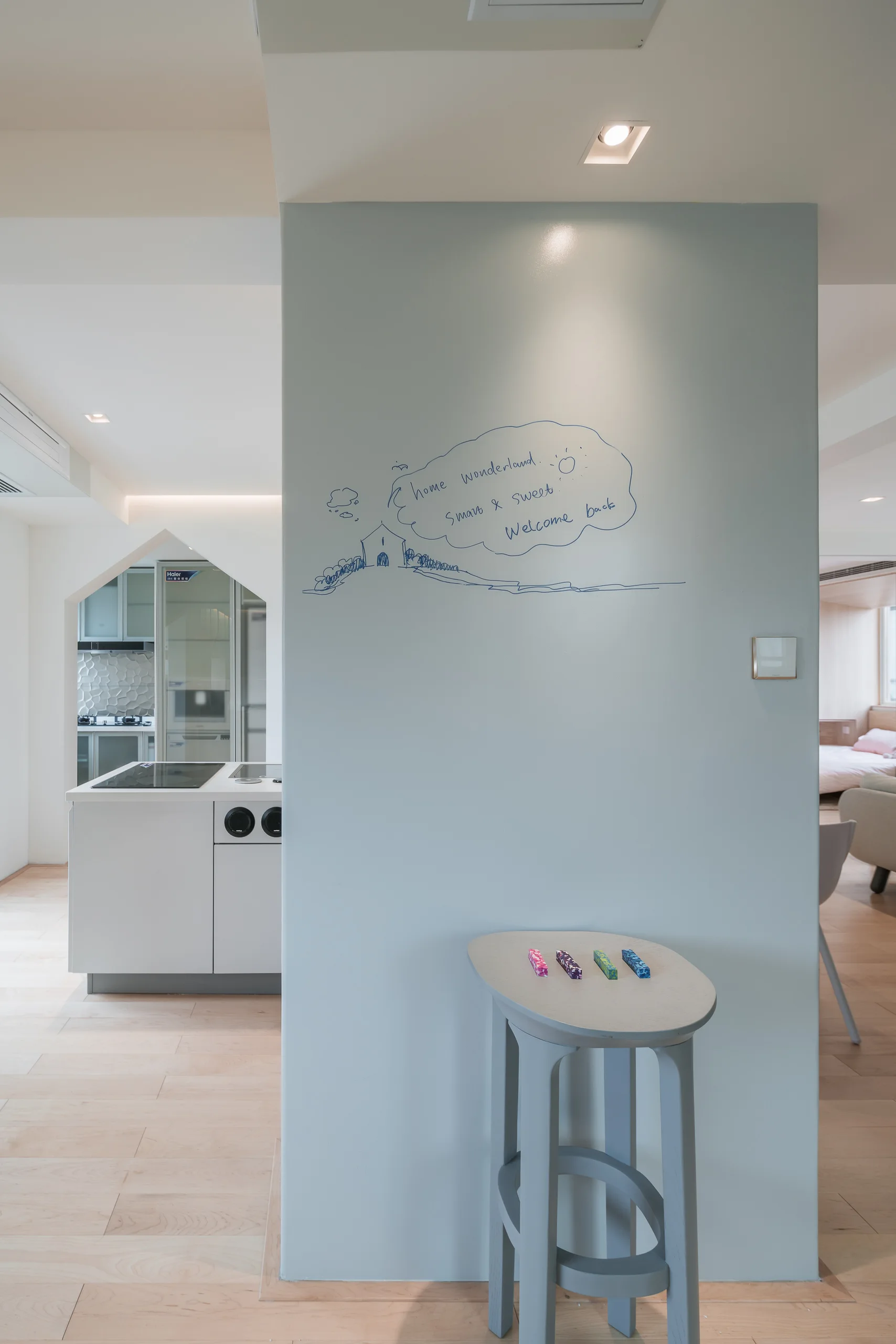 A light blue partition wall featuring a hand-drawn illustration stands next to a house-shaped archway leading into a minimalist white kitchen.