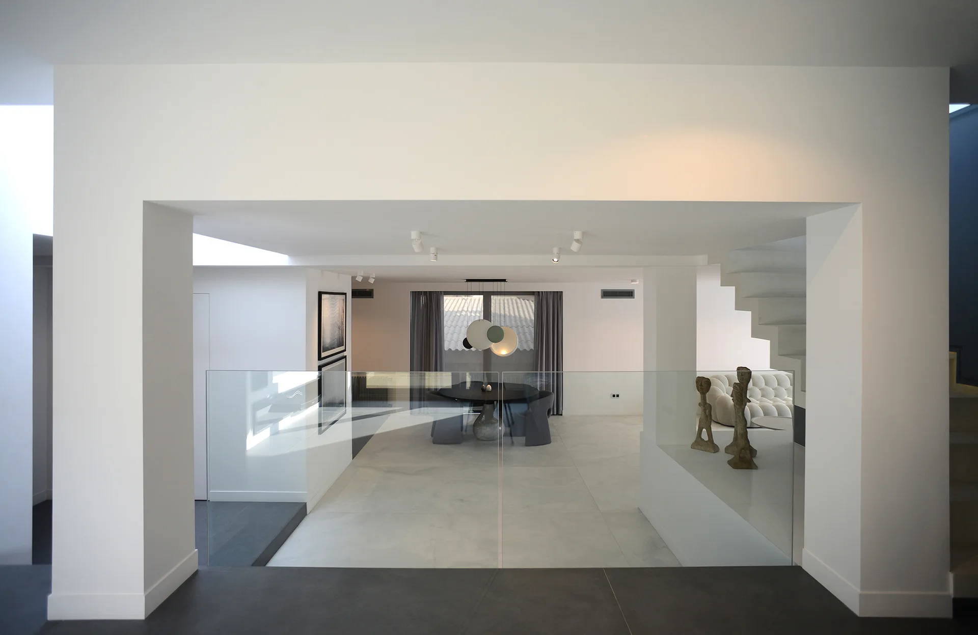 A view looking into a minimalist split-level interior featuring white walls, frameless glass railings, a black dining table, and a white staircase.