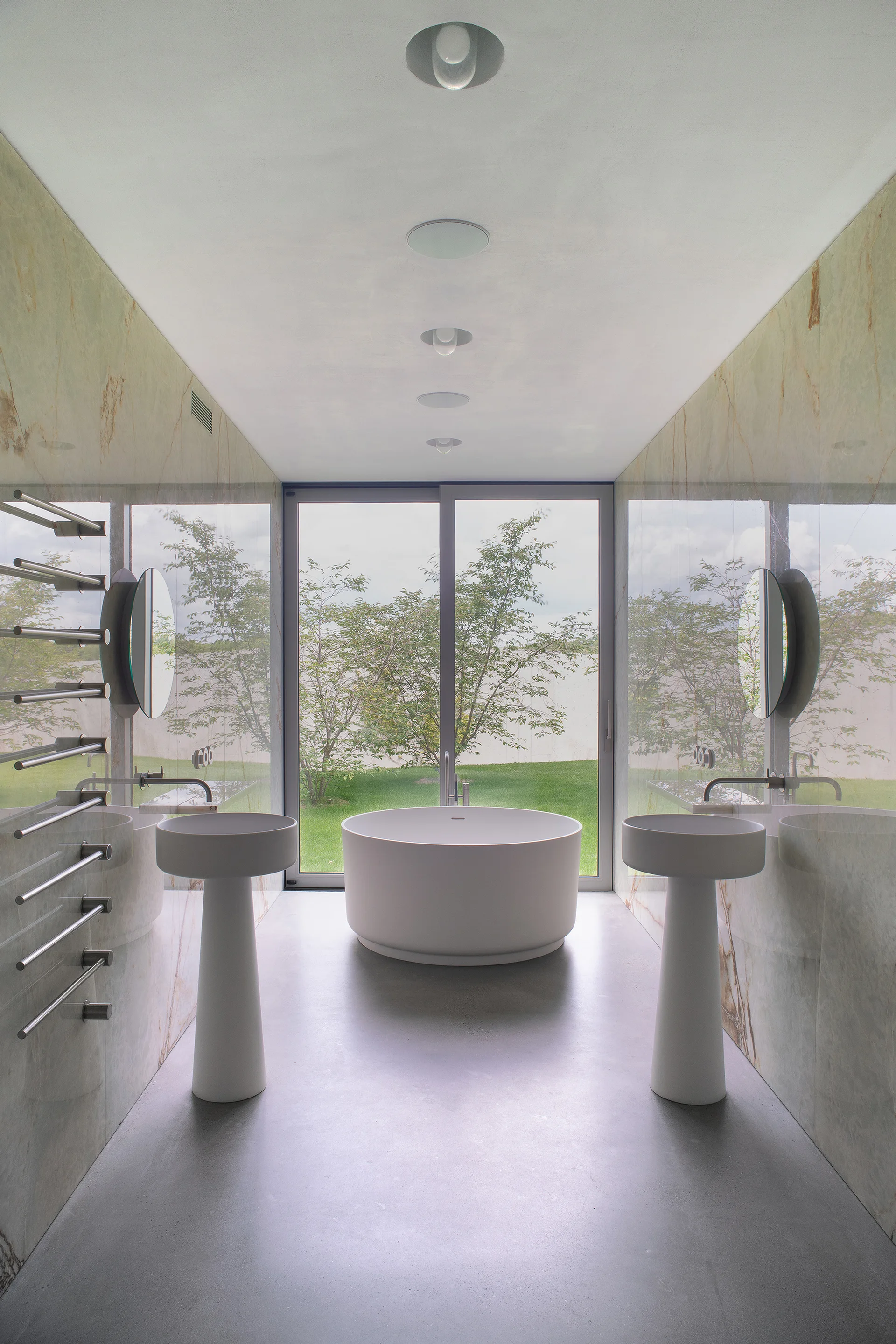 A symmetrical modern bathroom features a central circular soaking tub, two pedestal sinks against marble walls, and a large window facing a garden.