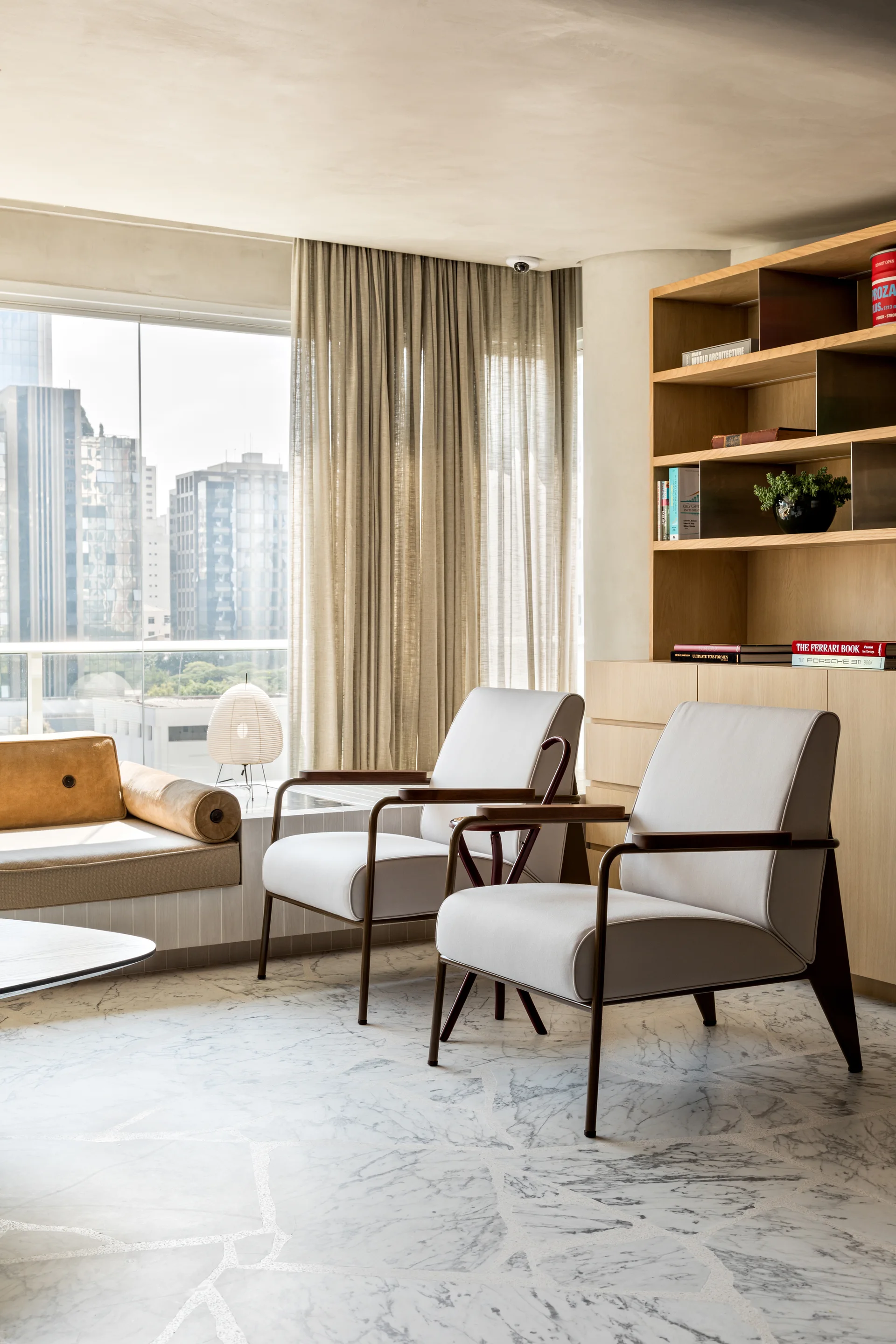 A bright living room features two light gray mid-century armchairs, a large wooden built-in shelving unit, and polished marble tiled flooring.