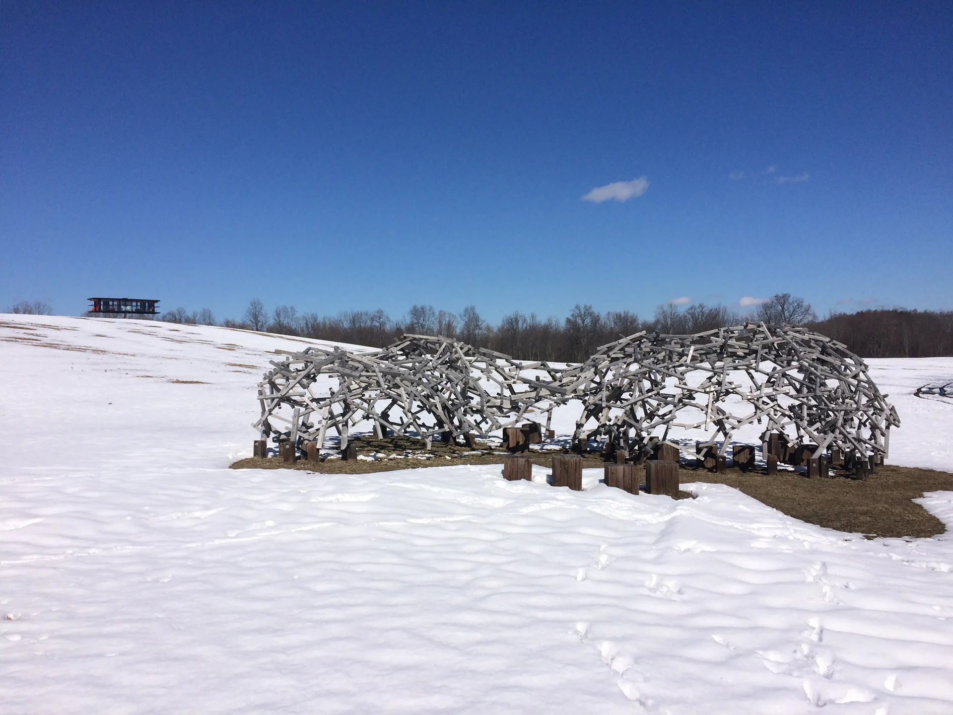 A large weathered wooden lattice sculpture stands in a snowy field under a clear blue sky.