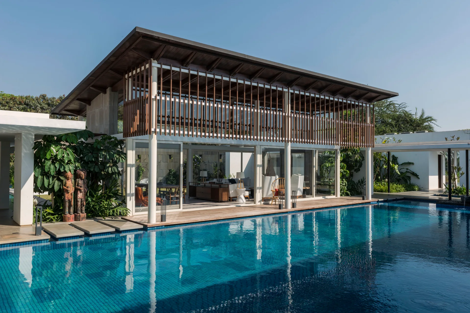 A contemporary house featuring sliding glass doors that open to a wood pool deck, a large swimming pool, and a vertical wood slatted balcony screen.