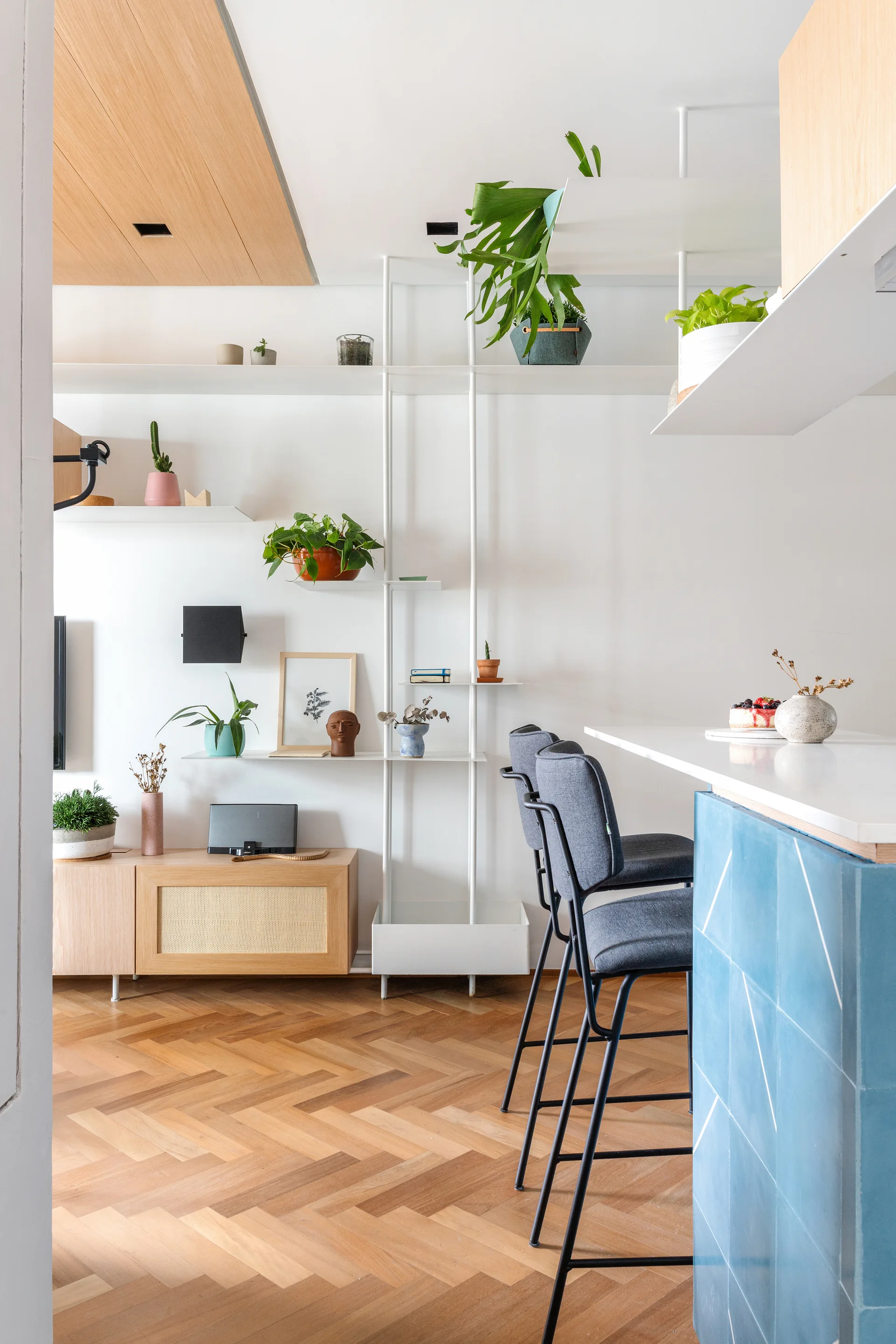 A modern kitchen area features a glossy blue tiled island with gray bar stools and minimalist white shelving holding potted plants.