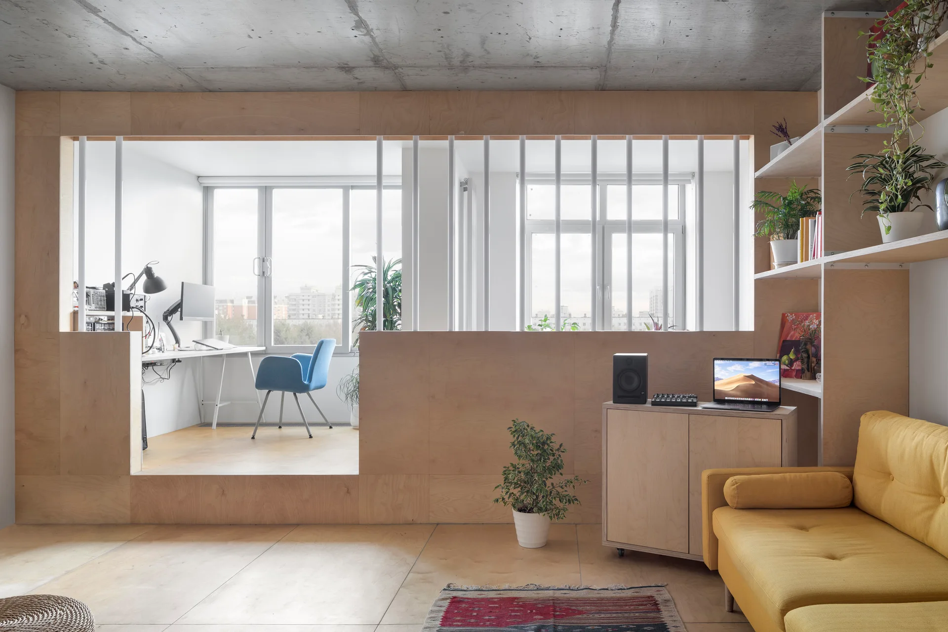 A modern living room with a yellow sofa, an exposed concrete ceiling, and a plywood partition leading to a raised home office.