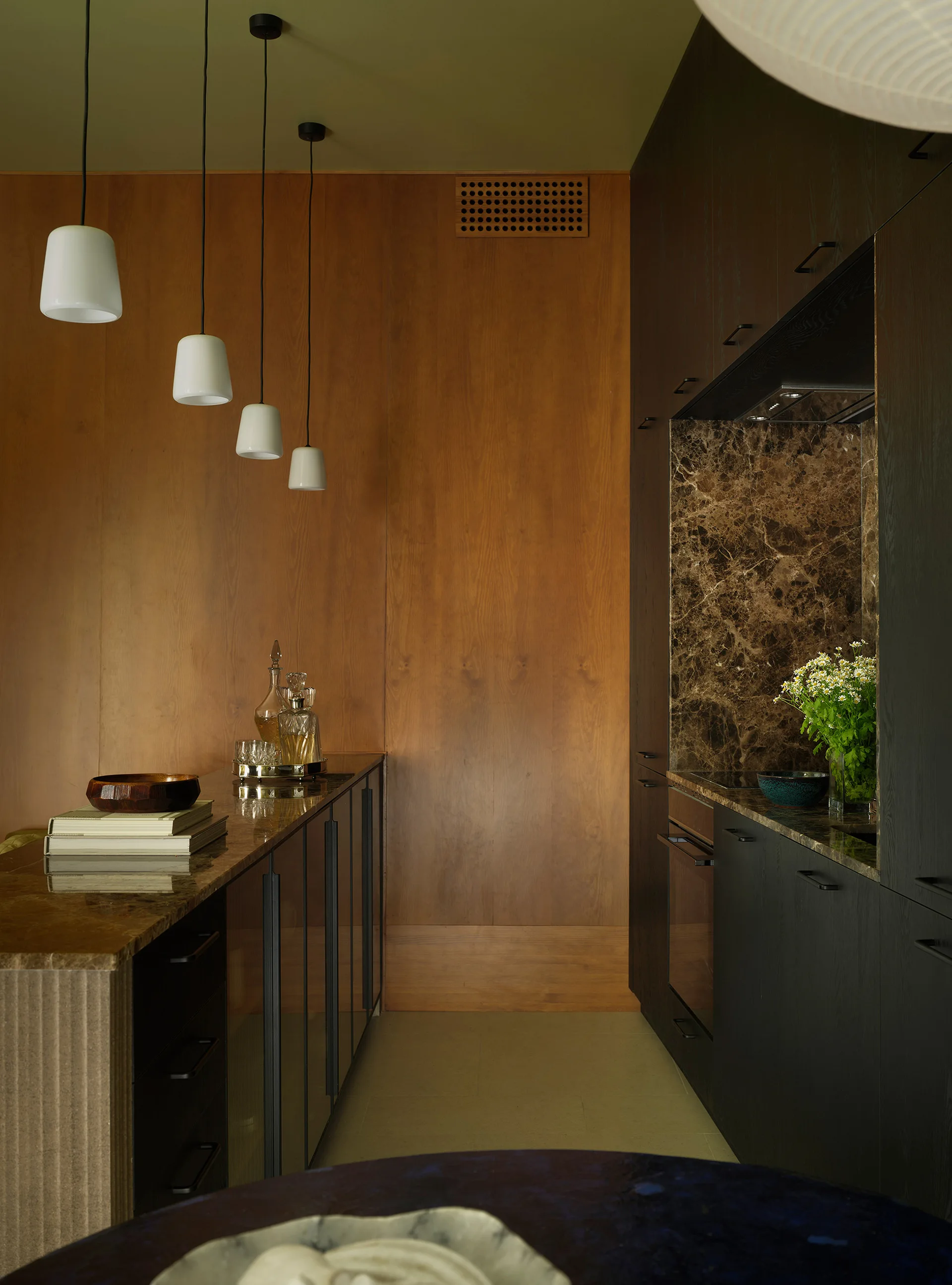 A galley kitchen featuring dark wood cabinets, a brown marble backsplash, a wood-paneled wall, and a row of white pendant lights.