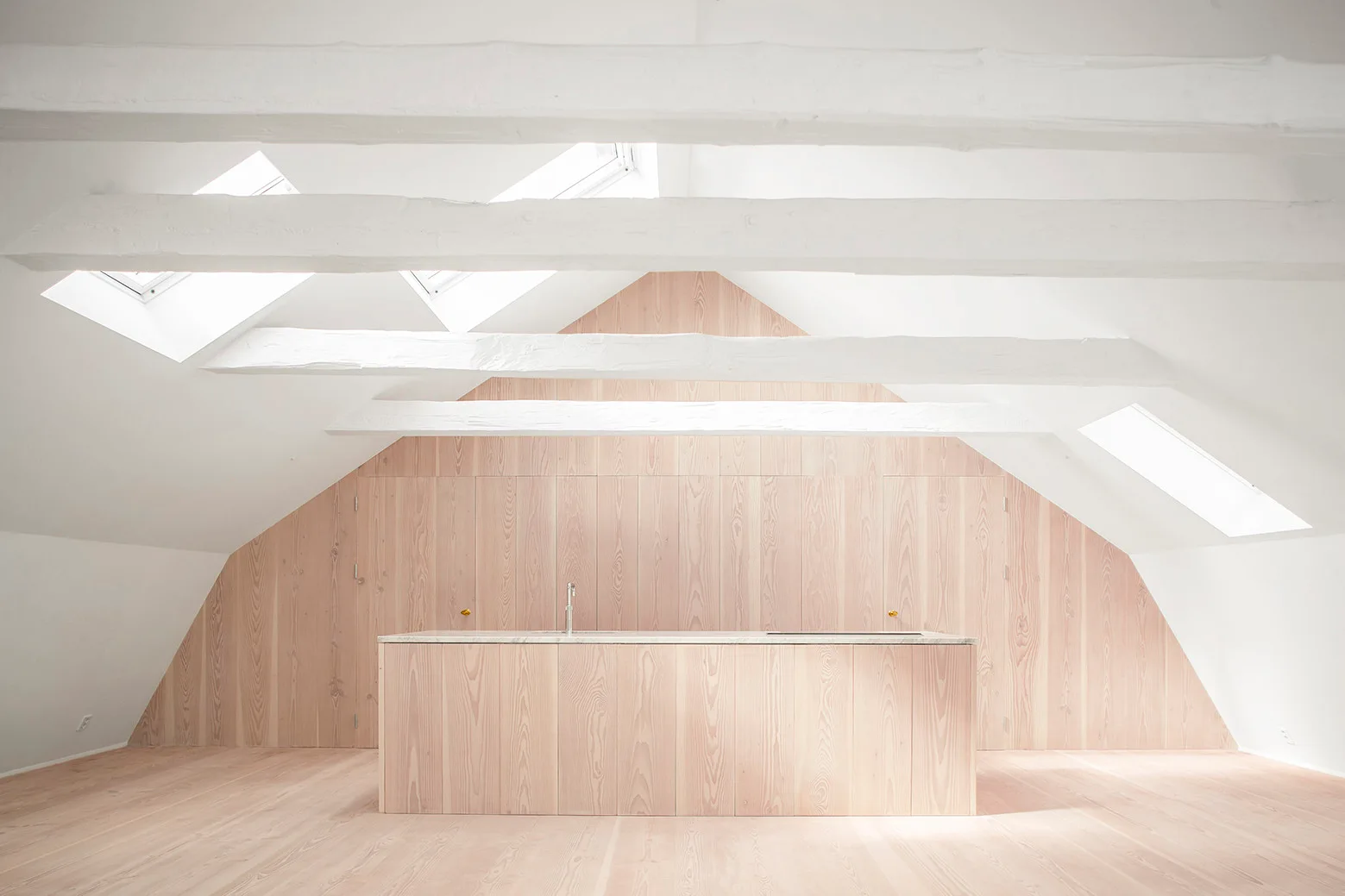 A minimalist attic kitchen features a matching pale wood island and paneled wall under sloped ceilings with white painted beams and skylights.