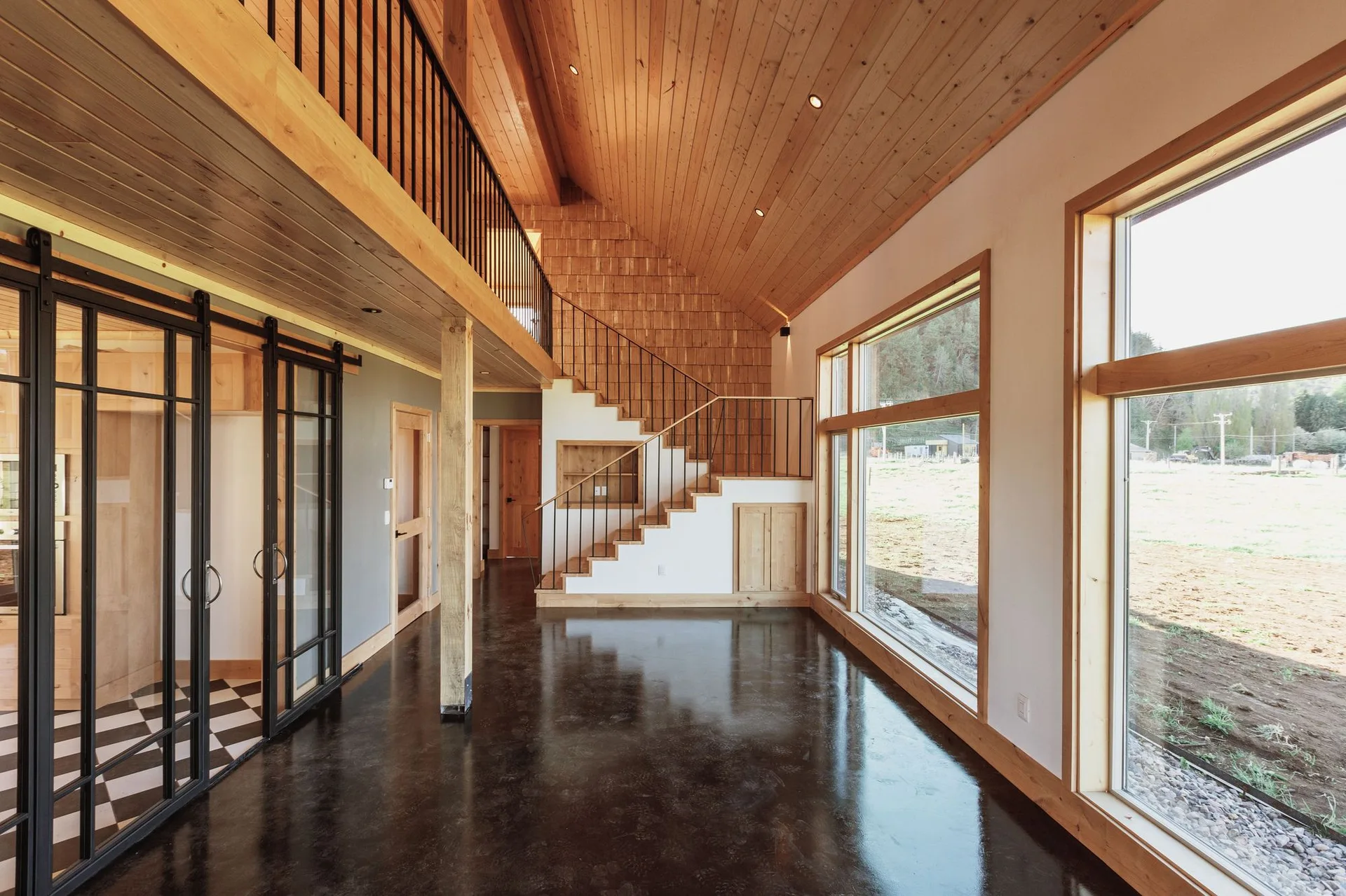 A modern rustic hallway features a vaulted wood ceiling, a staircase with black metal railings, and a reflective dark polished concrete floor.