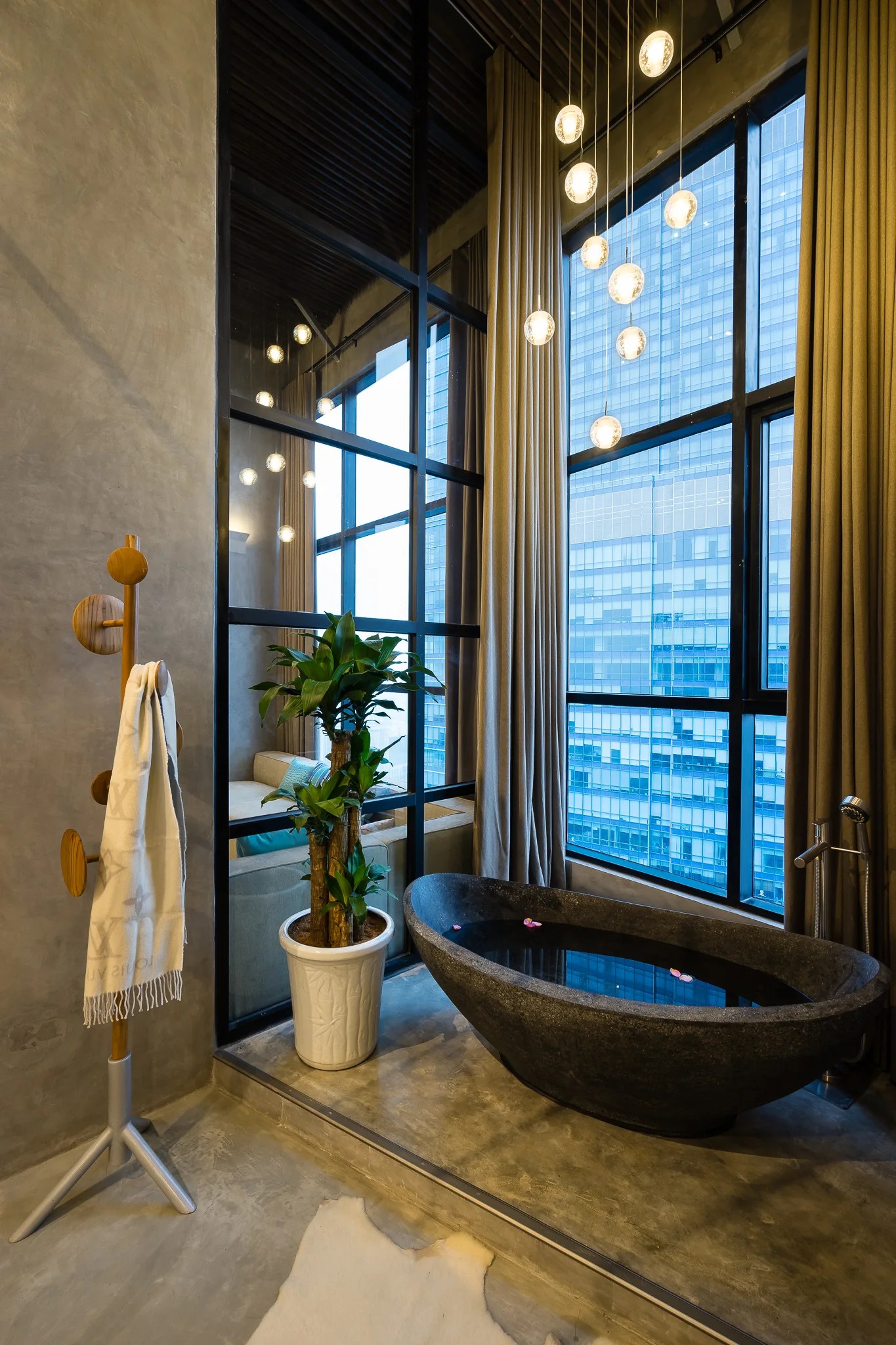A modern bathroom features a dark freestanding stone tub beside a tall window under a cluster of glass orb pendant lights.