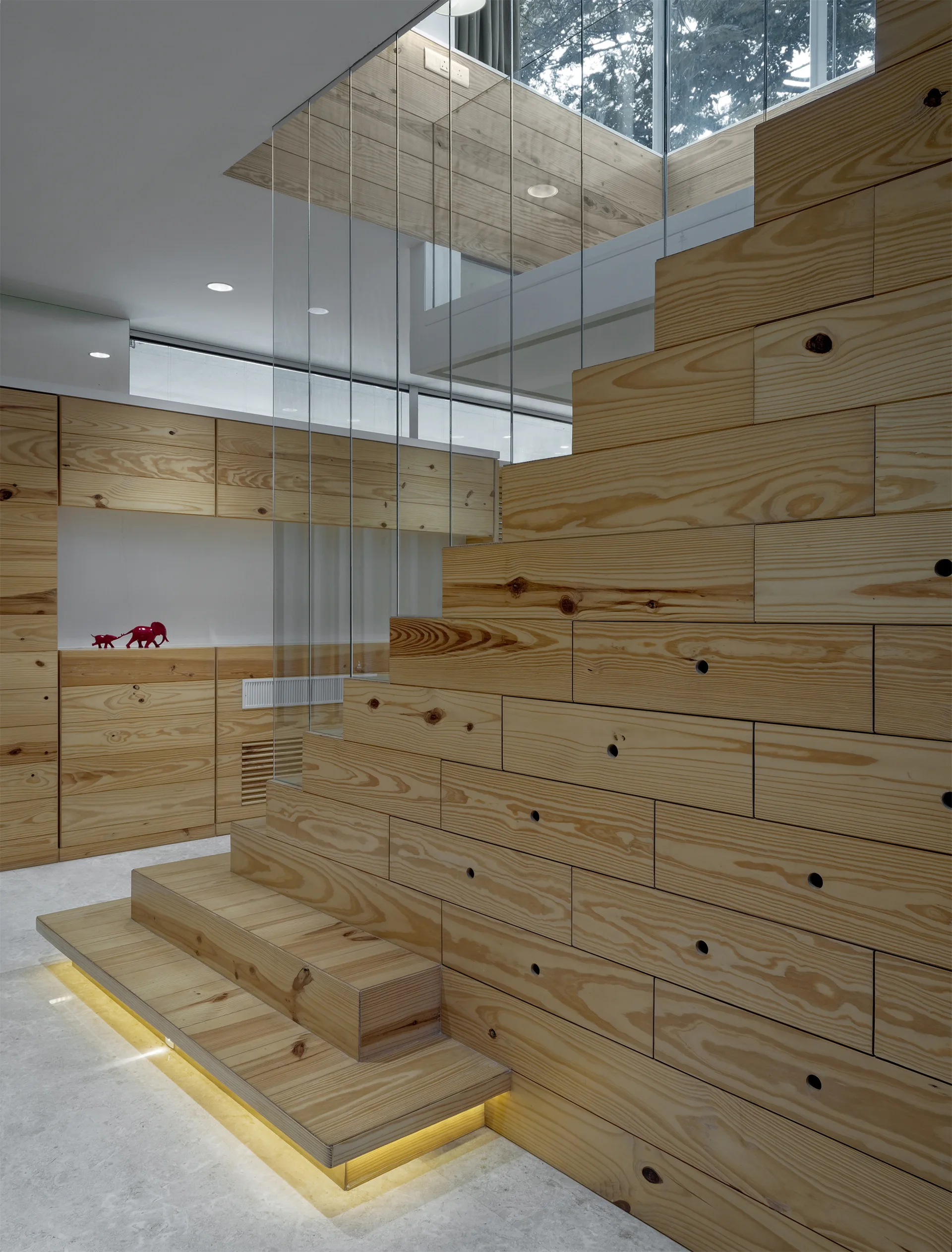 A modern pine wood staircase features integrated drawers on its side, a glass railing, and warm accent lighting underneath the bottom step.