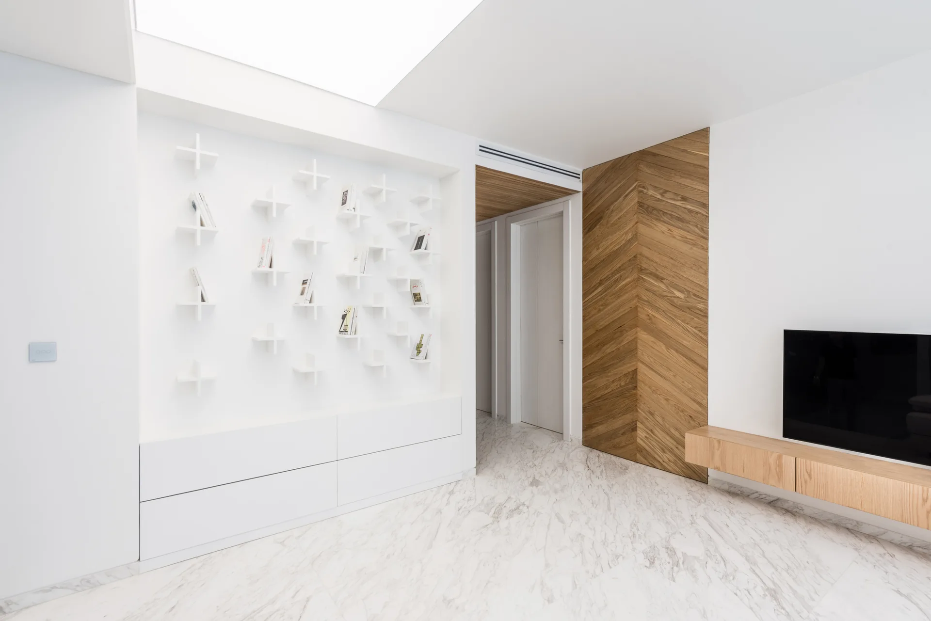 A minimalist white living space with custom cross-shaped wall shelves, a chevron wood accent wall, marble floor, and a skylight.