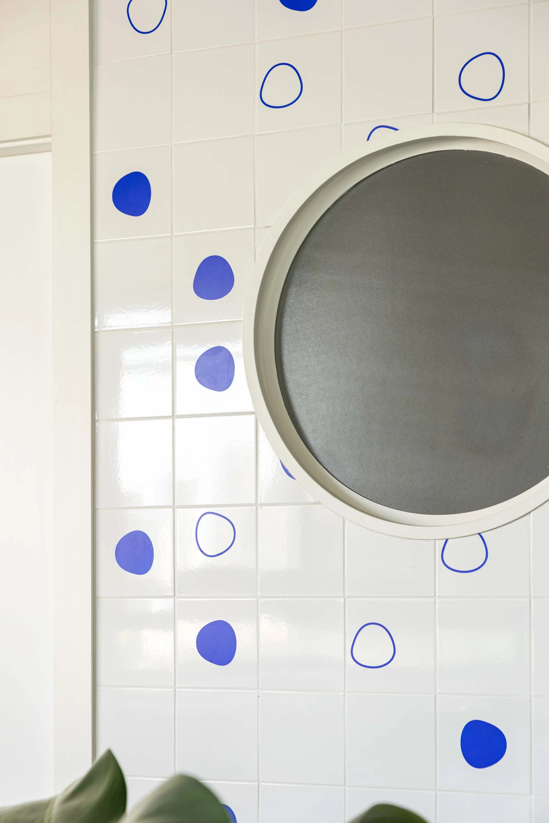 A close-up of a glossy white tiled wall decorated with cobalt blue polka dots and solid circles, featuring a large round mirror.