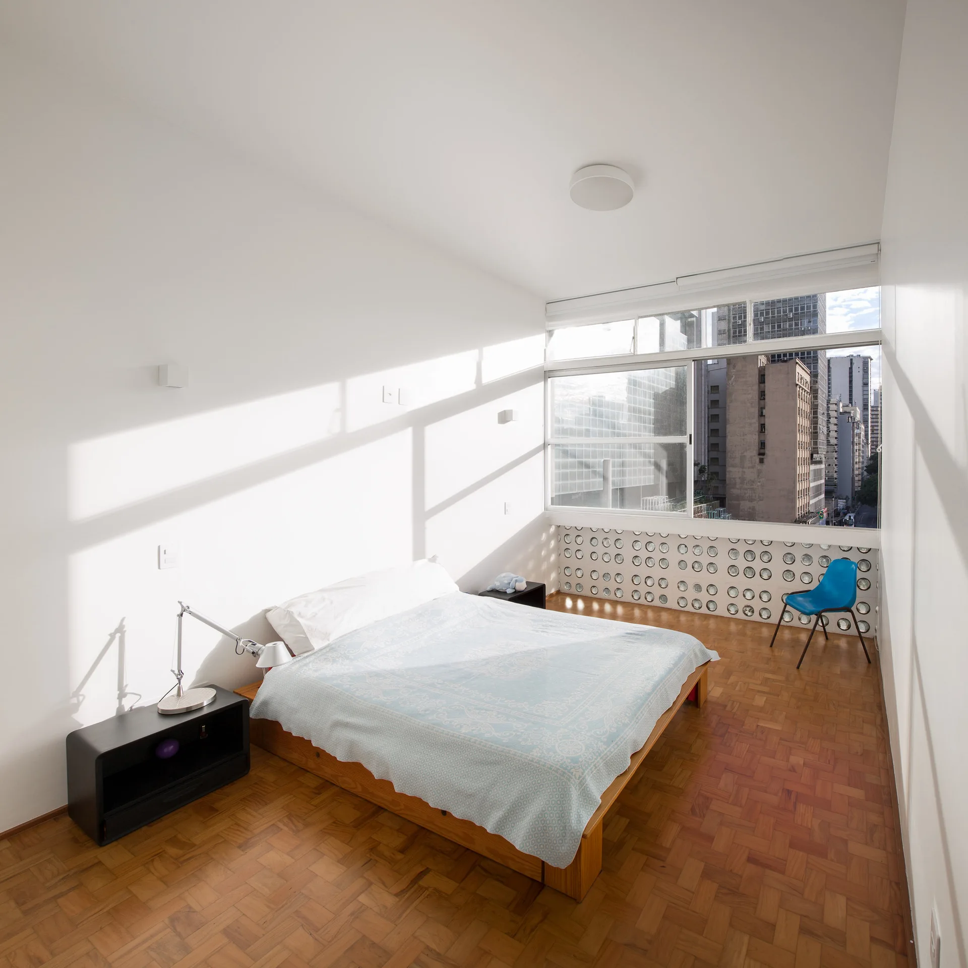 A minimalist bedroom featuring parquet wood flooring, a platform bed, a wide window, and a circular glass block wall detail.