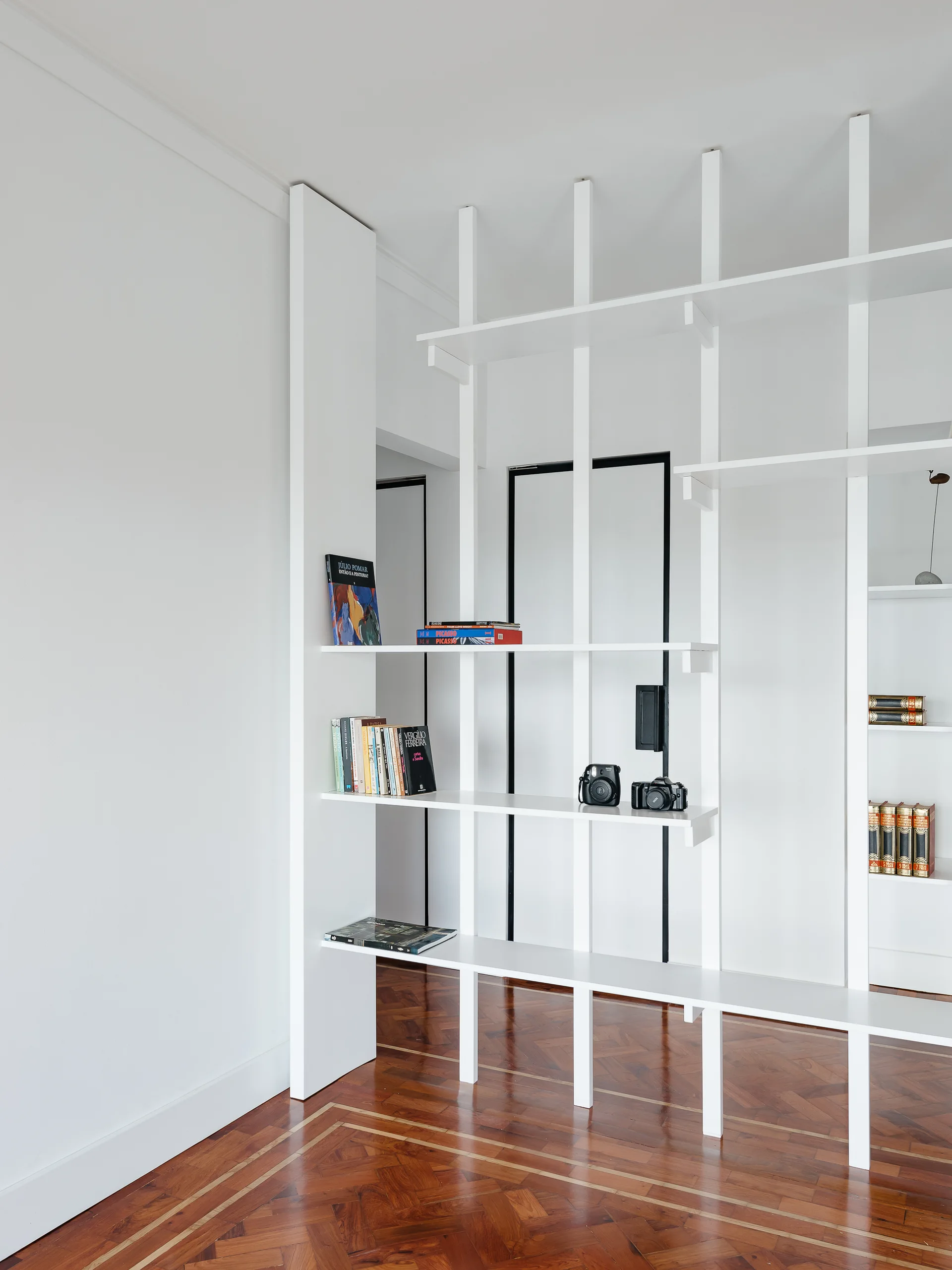 A custom floor-to-ceiling white open shelving unit serves as a partition over warm herringbone wood floors in a minimalist home.