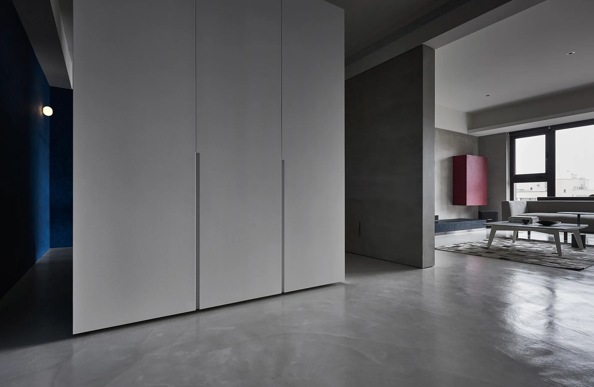 A minimalist room features polished concrete floors, a large white wardrobe partition, and a distant living area with a red cabinet and sofa.