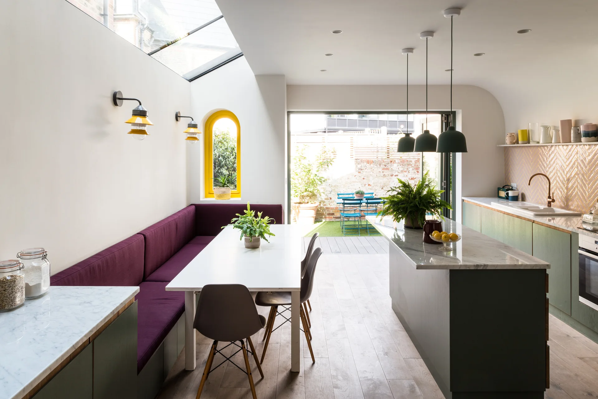 An open kitchen features a purple cushioned banquette, a white dining table, a marble-topped island, sage green cabinets, and a glass skylight.