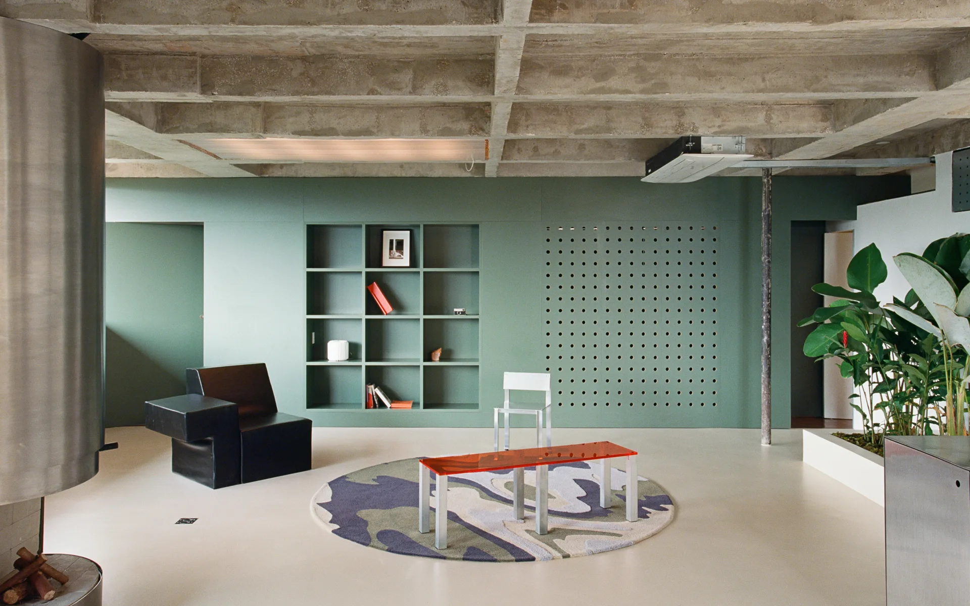 A minimalist living room features a sage green wall with built-in shelving, an exposed concrete ceiling grid, and a geometric black armchair.