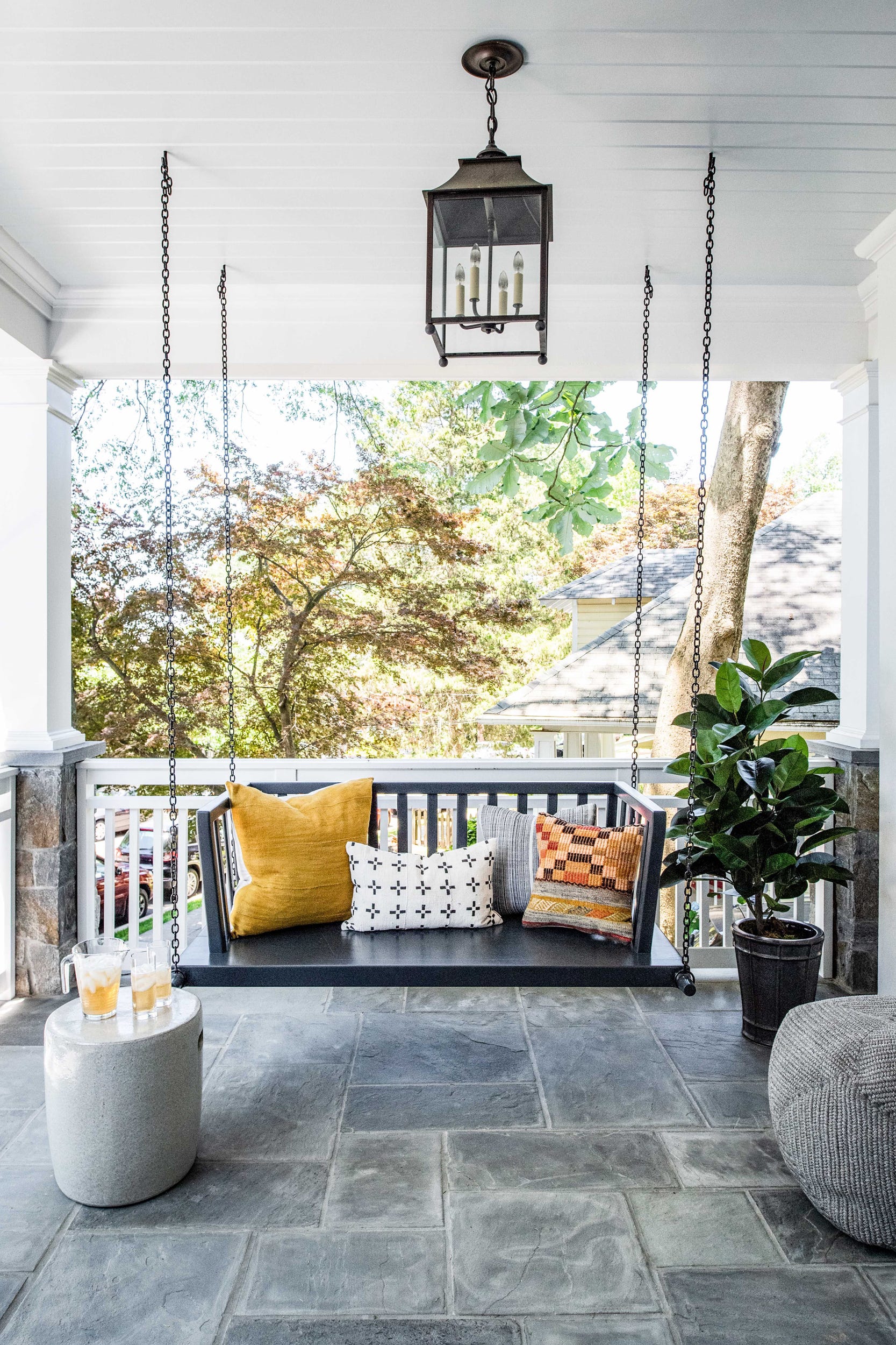 A black wooden hanging swing with patterned pillows sits on a stone-tiled porch with white columns and a hanging lantern.