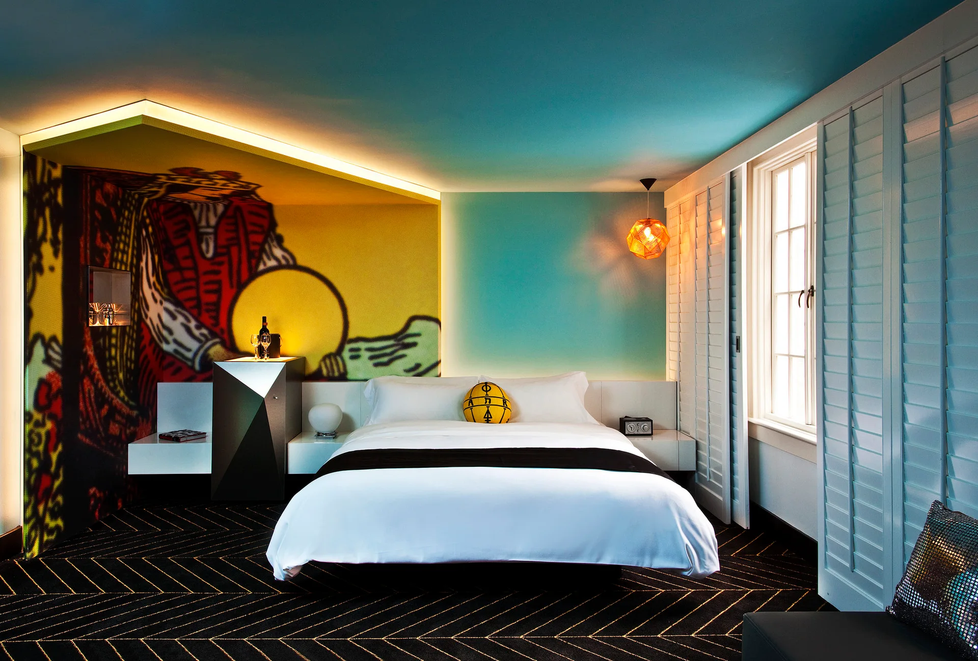 A modern bedroom features a teal ceiling, a large colorful pop art mural, white platform bedding, and a black and gold chevron carpet.