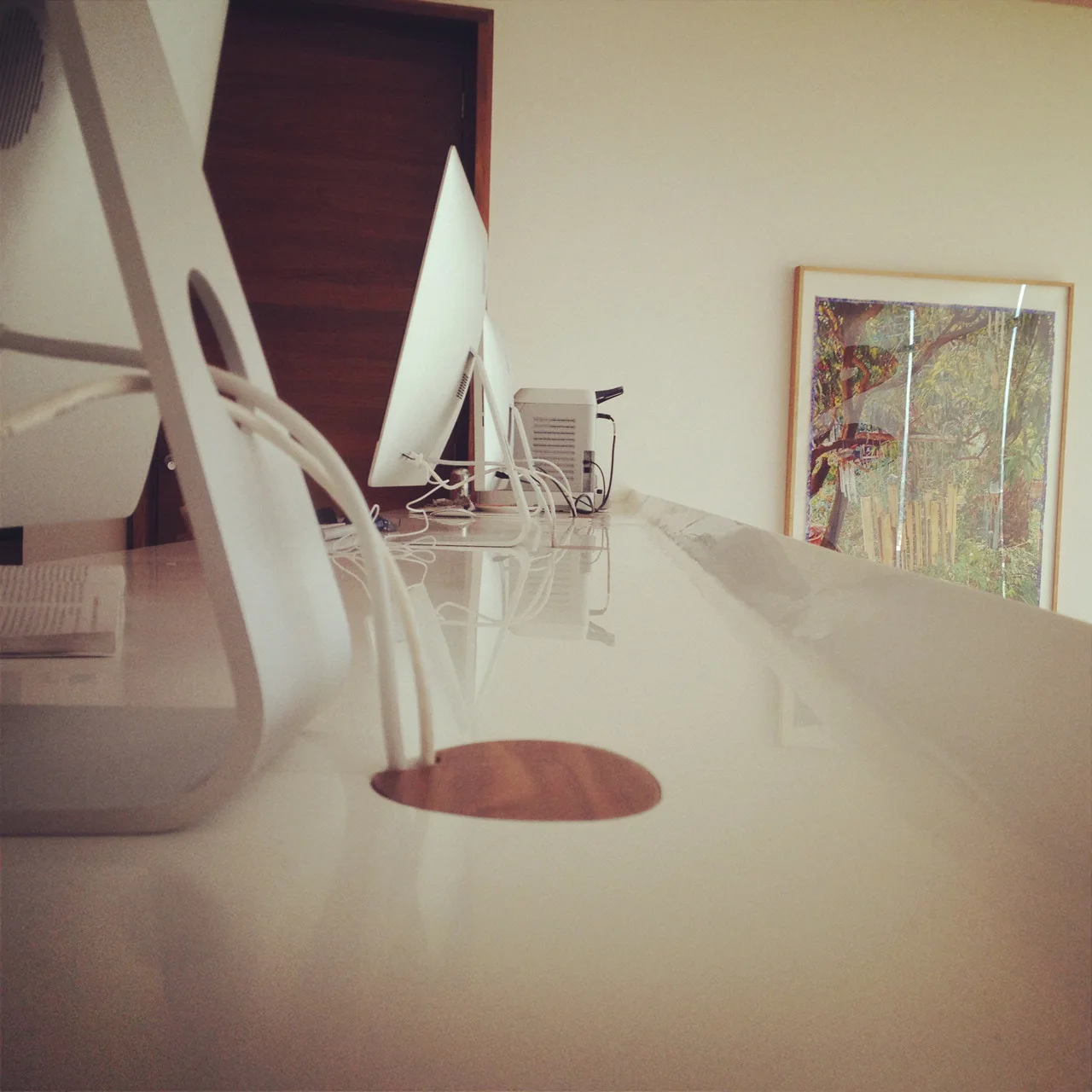 A close-up view of a long white glossy desk surface with computer monitors and a circular wood grommet for cable management.