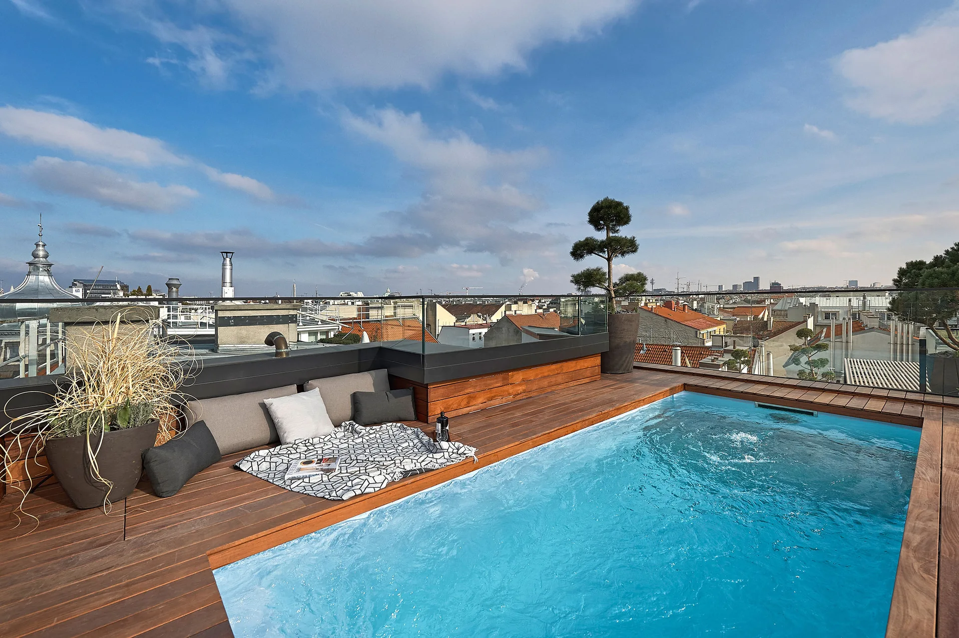 A rooftop swimming pool is surrounded by warm wood decking, cushioned outdoor seating, and a clear glass railing overlooking a city skyline.