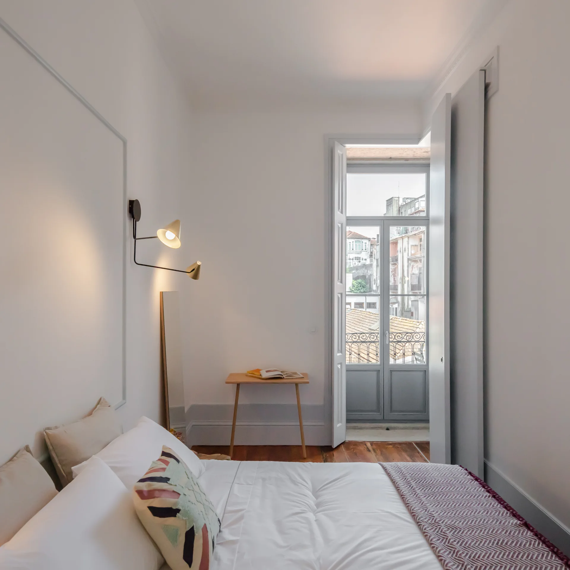A bright bedroom features a white bed, a small wooden desk, a black wall-mounted reading lamp, and tall glass doors opening to a balcony.