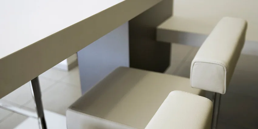 Two minimalist cream upholstered bar stools with low backrests are tucked under a thick cream kitchen countertop.