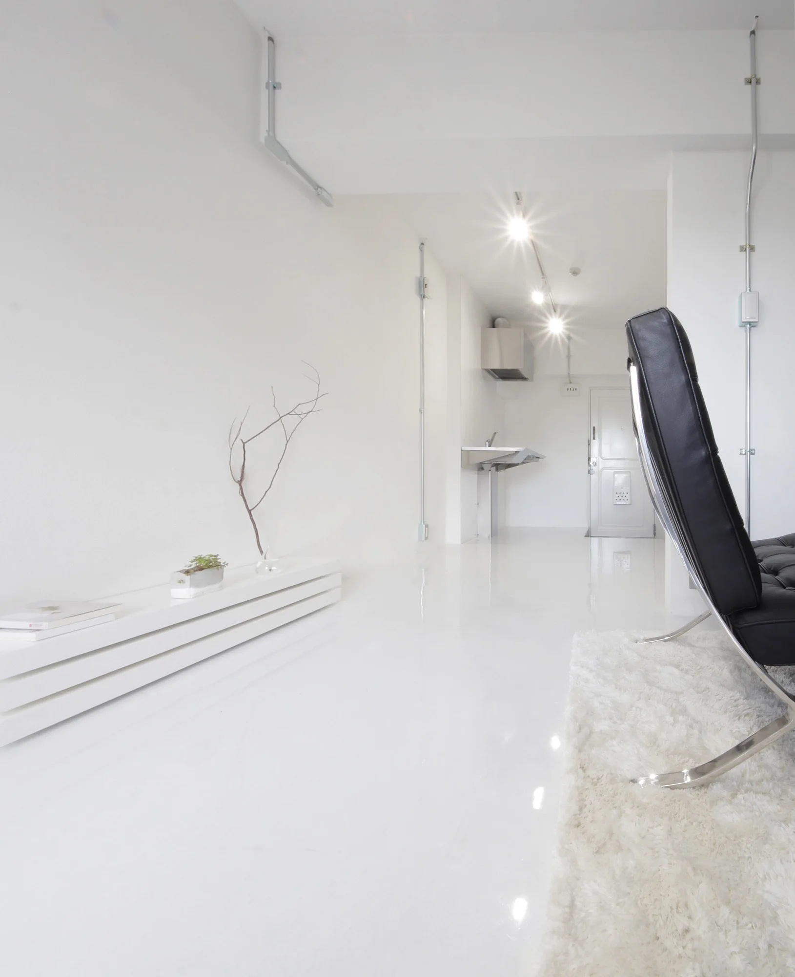 A stark white minimalist living space features a black leather lounge chair on a white shag rug, glossy white floors, and exposed wall conduits.