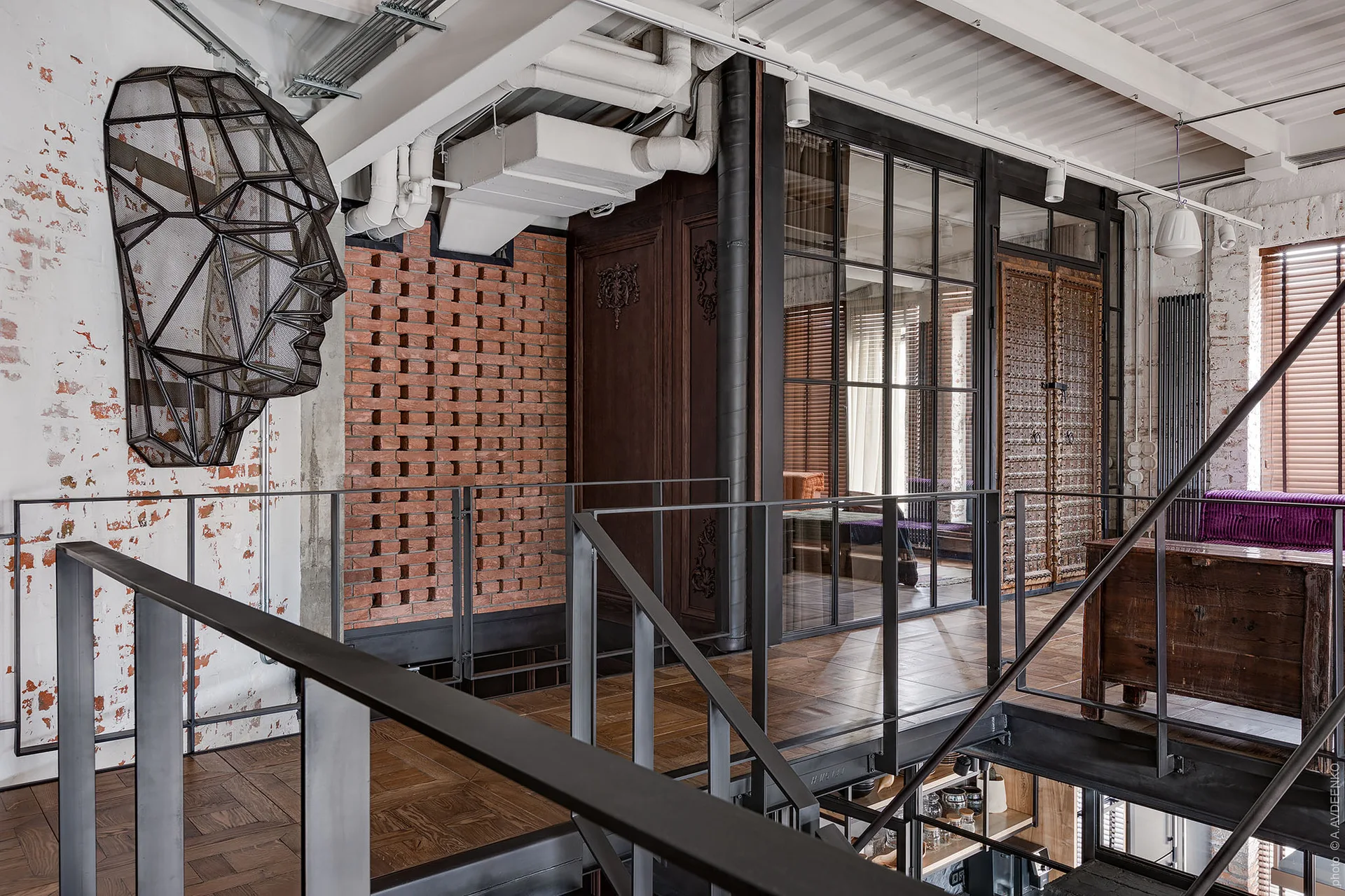 An industrial loft landing features a black metal staircase, a wire mesh face sculpture on a brick wall, and a black-framed glass partition.