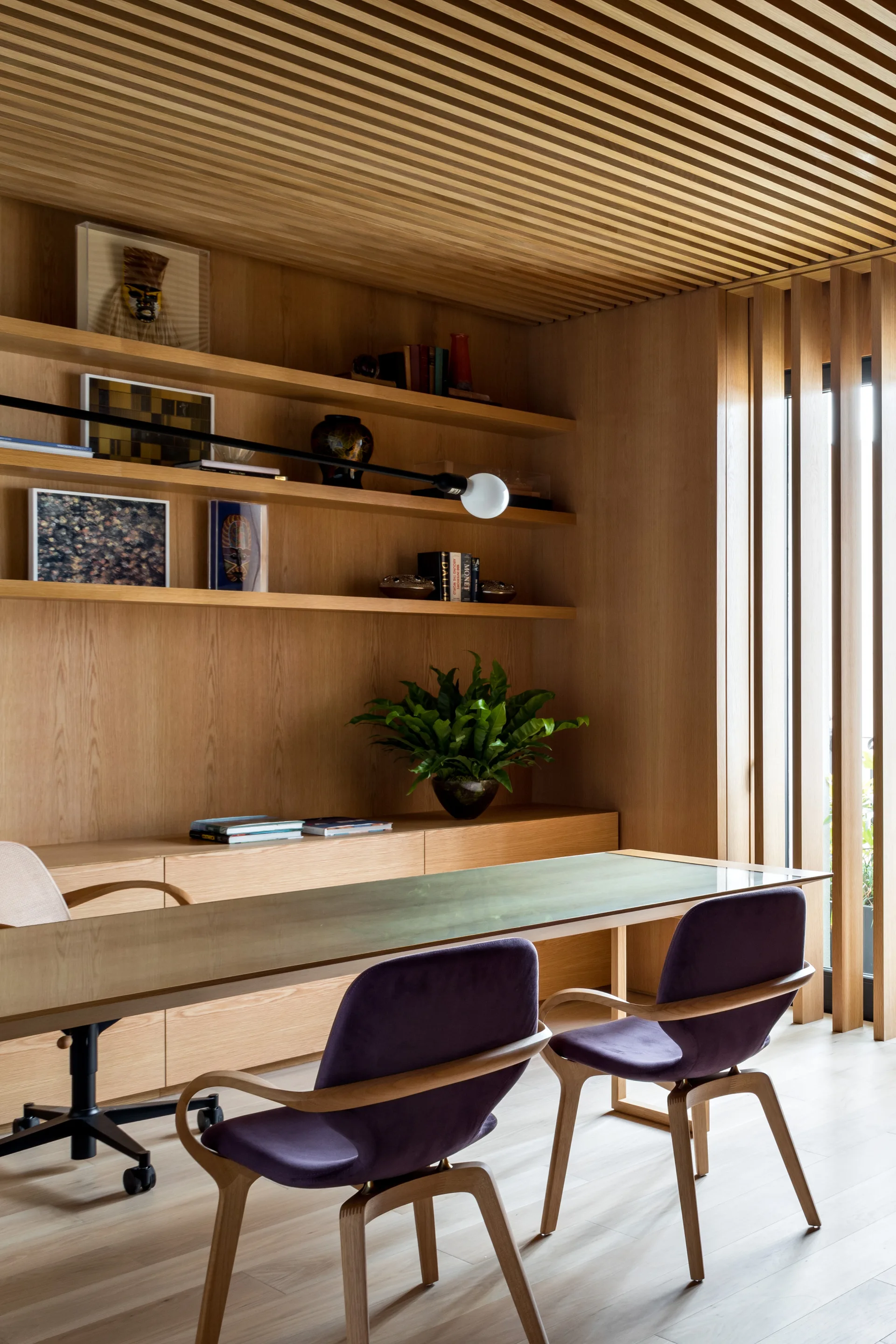 A modern home office with built-in light wood cabinetry, a glass desk, and two purple armchairs under a slatted ceiling.