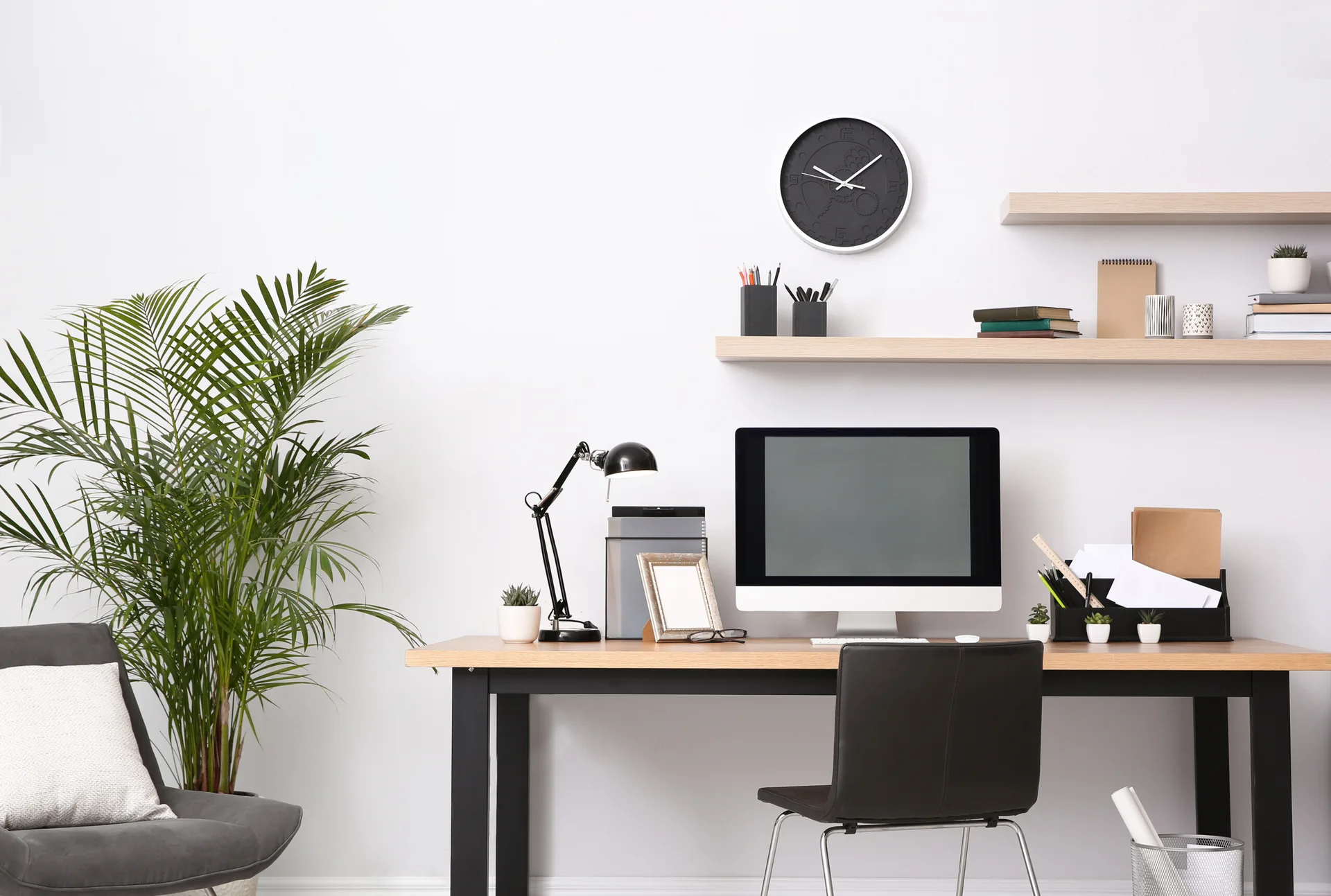 A wooden writing desk with a desktop computer and black chair sits under two light oak floating shelves on a white wall.