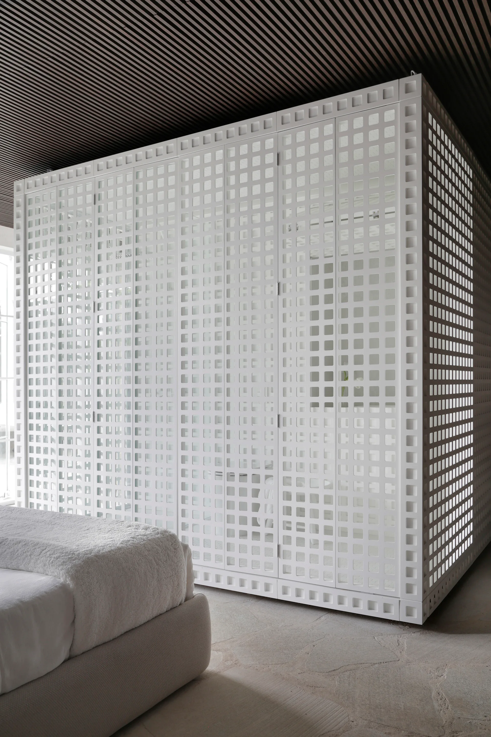 A modern bedroom features a large white wardrobe with a square grid pattern, set next to a textured white bed below a dark slatted ceiling.