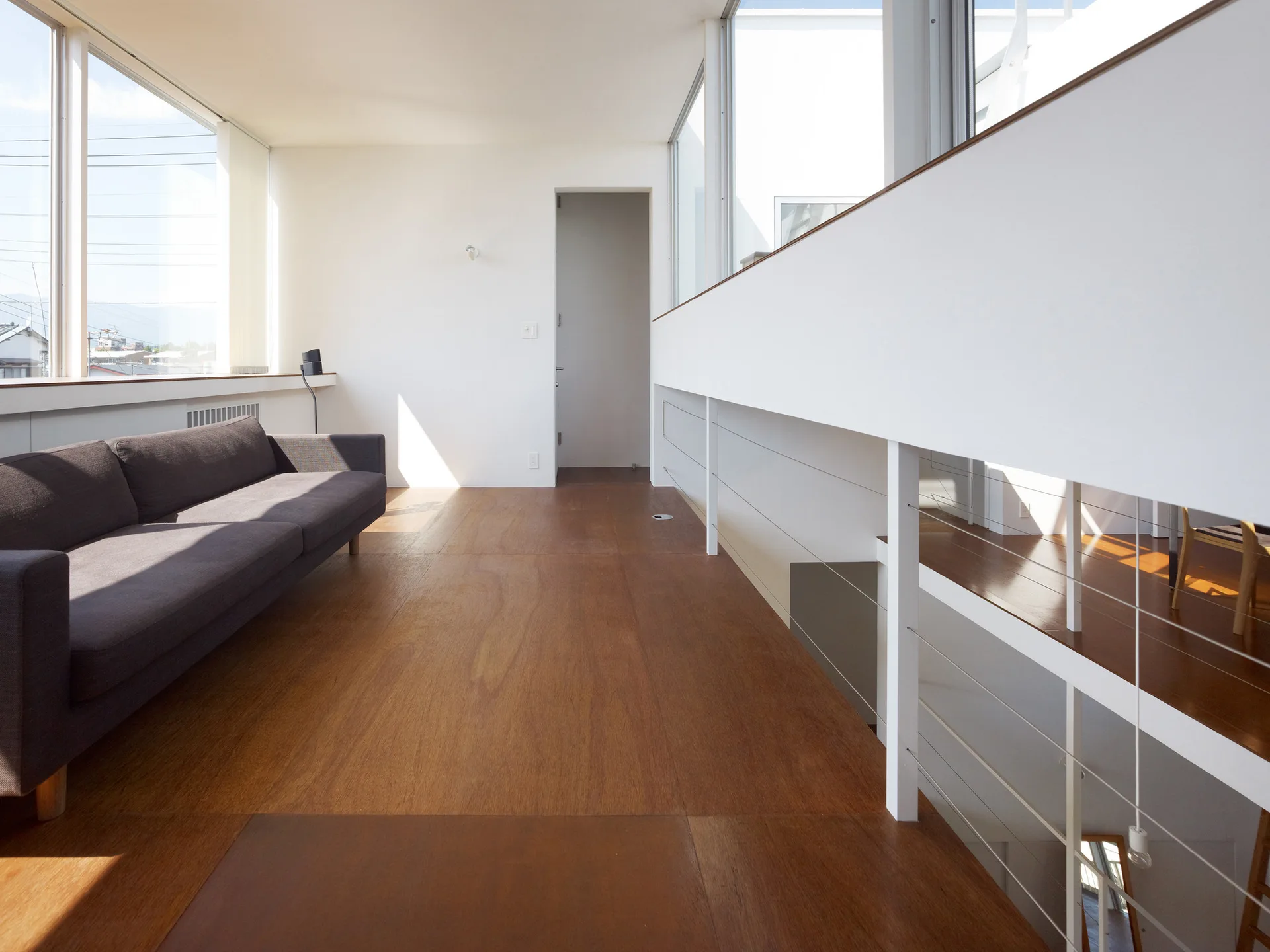 A minimalist mezzanine living area features a dark gray sofa on warm wood floors next to a white metal staircase railing and large windows.