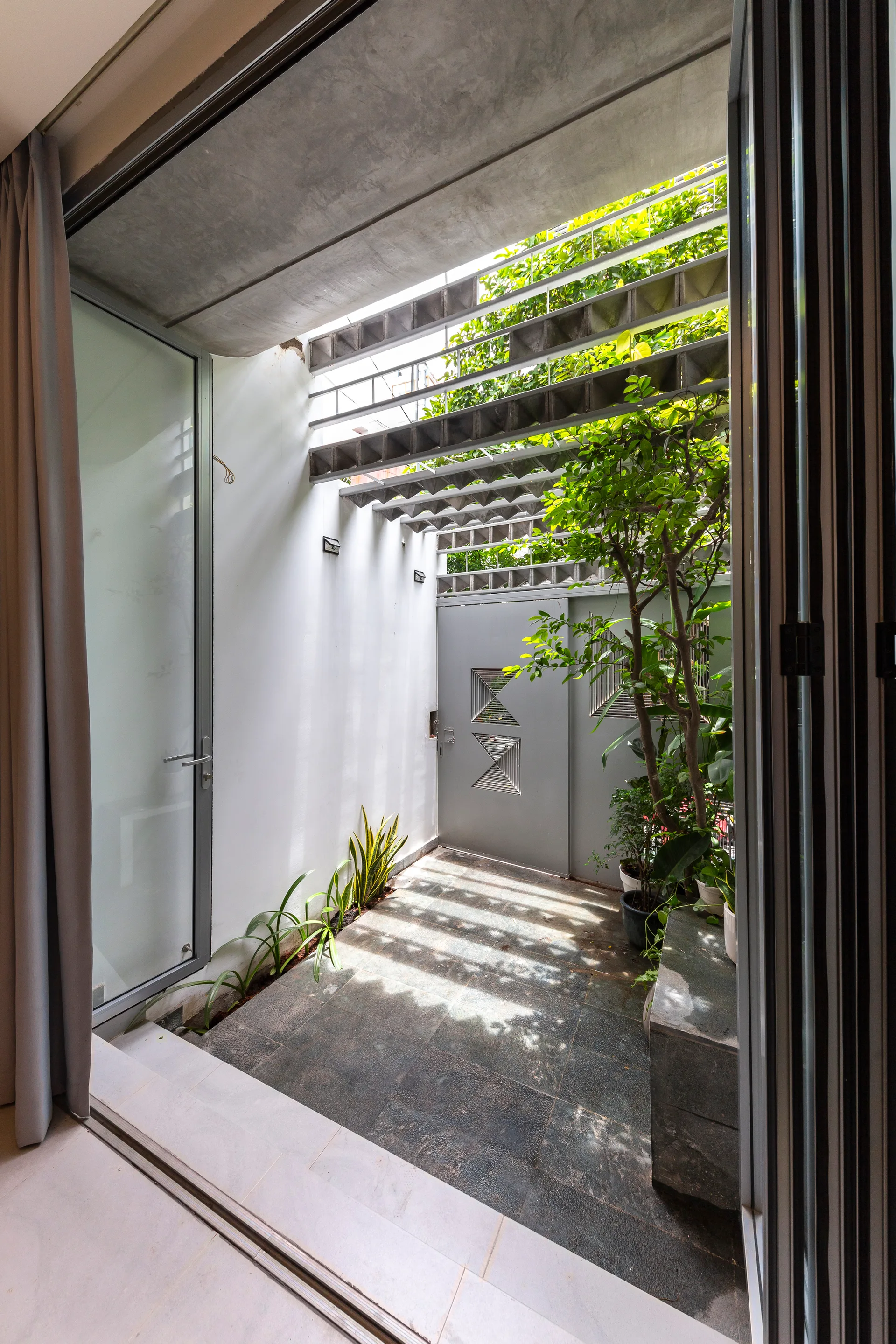 An outdoor courtyard viewed through sliding glass doors, featuring dark stone flooring, white walls, green potted plants, and a slatted metal ceiling.