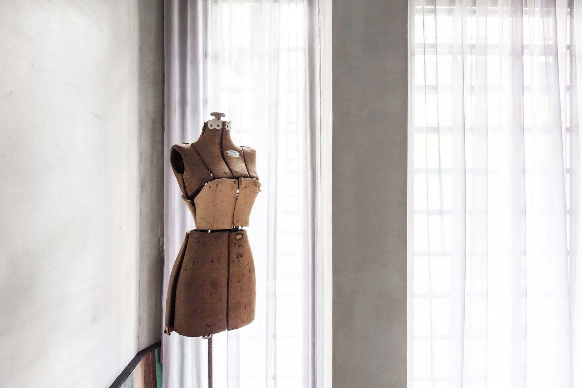 A vintage tan dress form stands in front of tall windows covered with sheer white curtains next to a grey concrete wall.