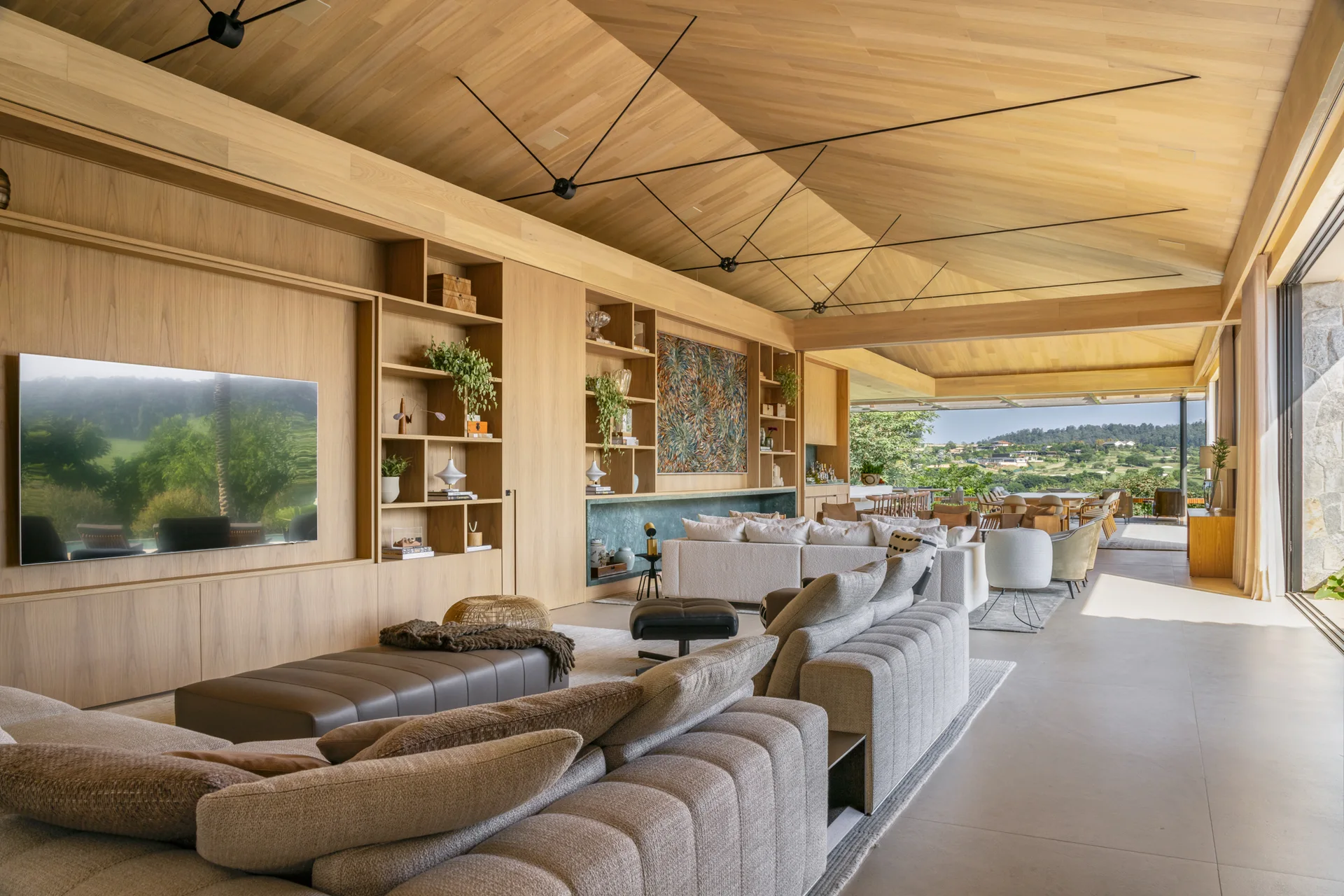 A spacious open-plan living room features modular sofas, an integrated wood wall with built-in shelves, a vaulted ceiling, and sliding glass doors.