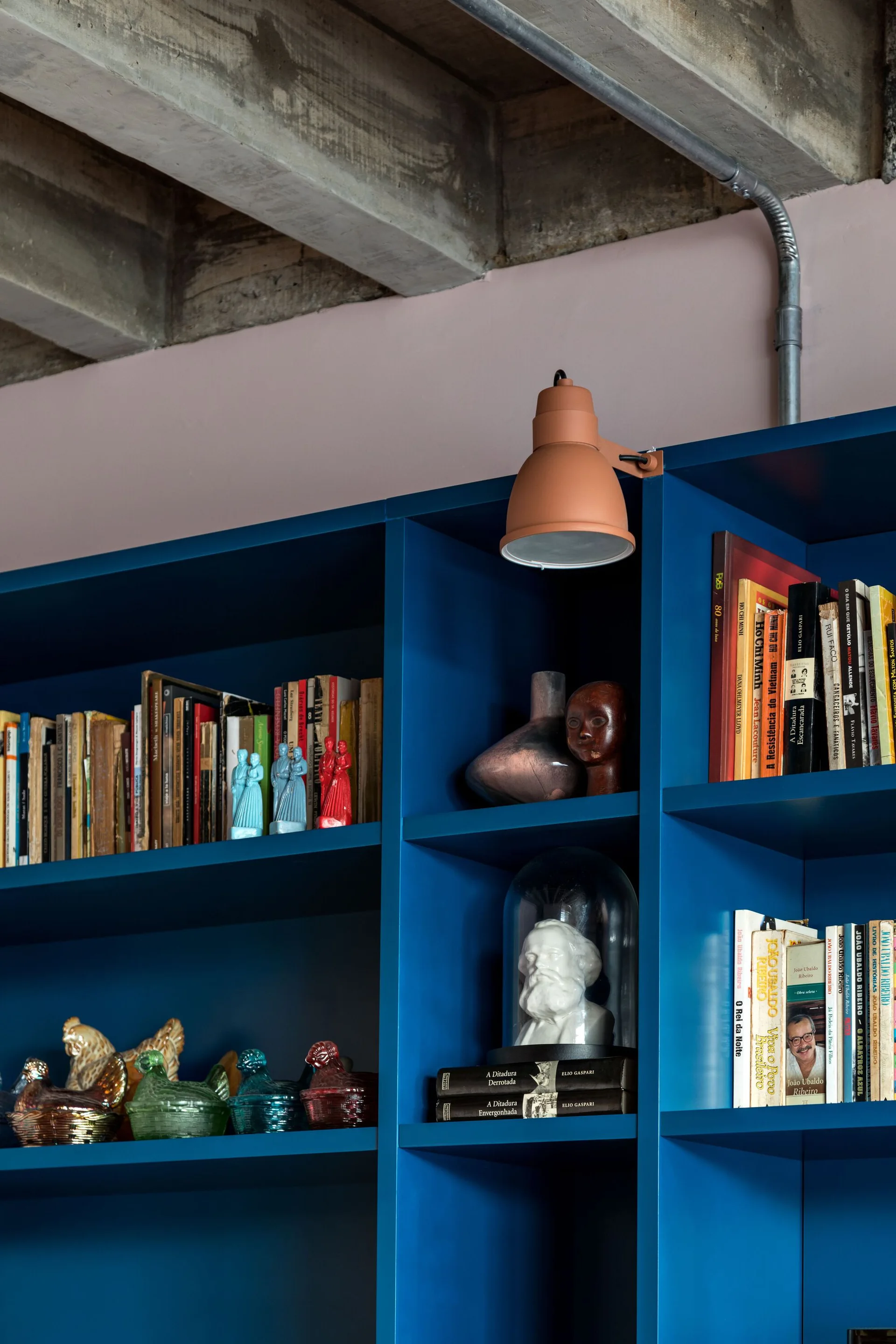 A saturated blue bookshelf filled with books and ceramic decor stands beneath an exposed concrete ceiling with a terracotta wall lamp attached.