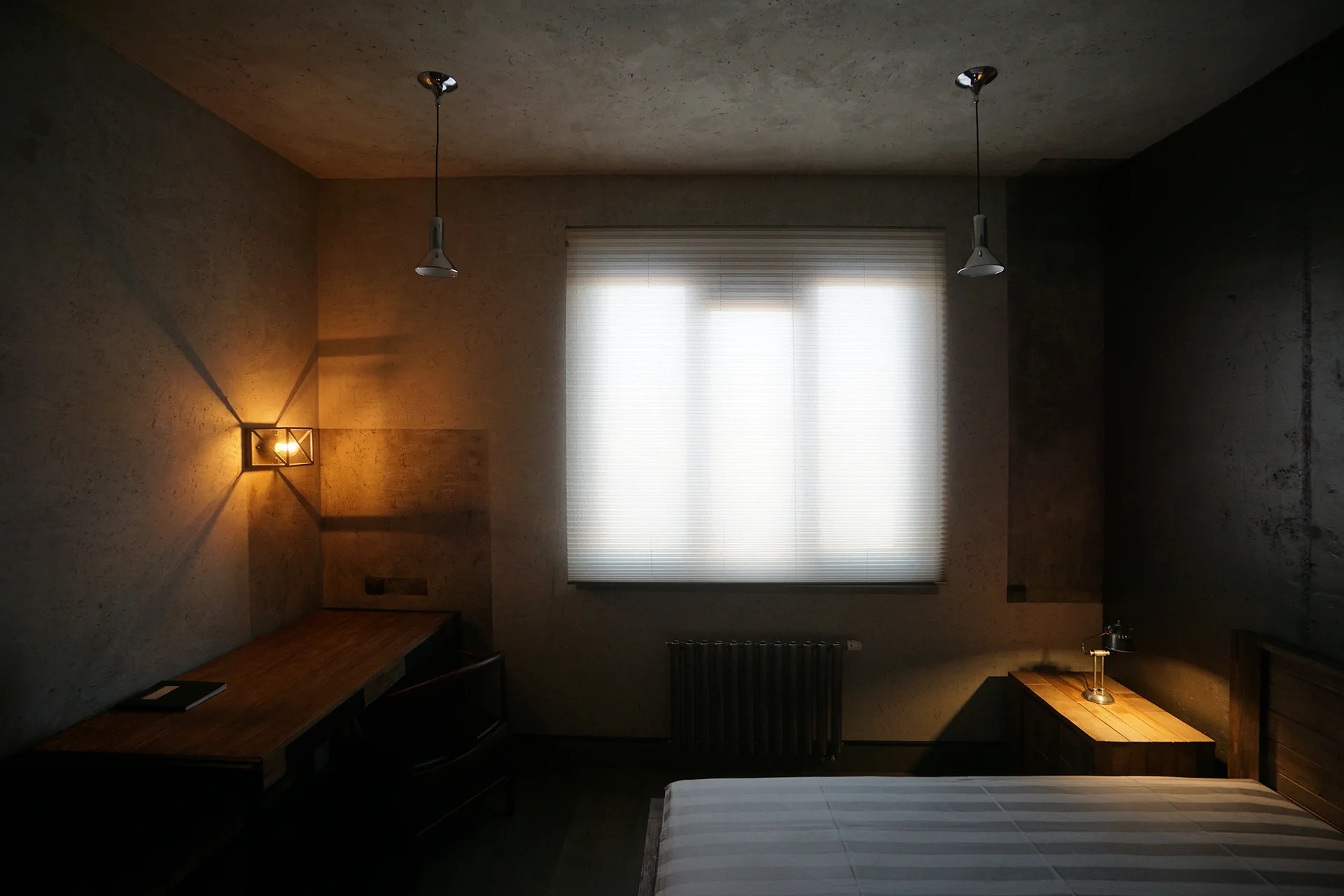A moody grey industrial bedroom with concrete-textured walls, a wooden desk under a warm wall sconce, and a window with white blinds.