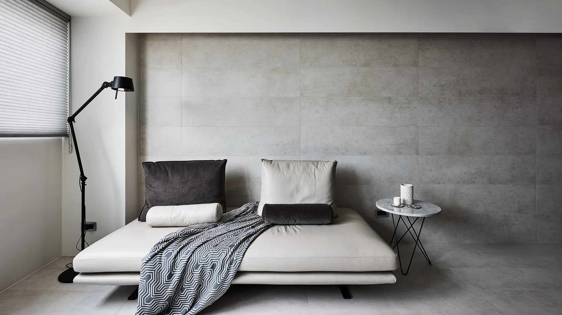 A minimalist leather daybed with a geometric throw blanket sits against a grey tile wall next to a black floor lamp and a marble side table.